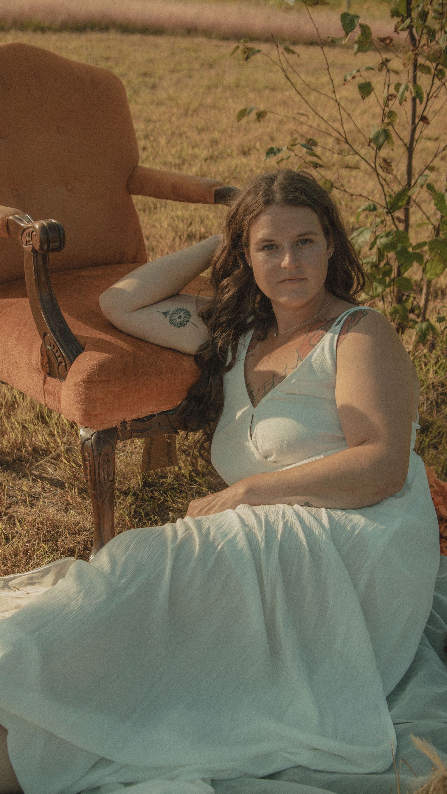Figure in a long turquoise dress lies gracefully outdoors against a rustic brown backdrop near greenery in warm sunlight.