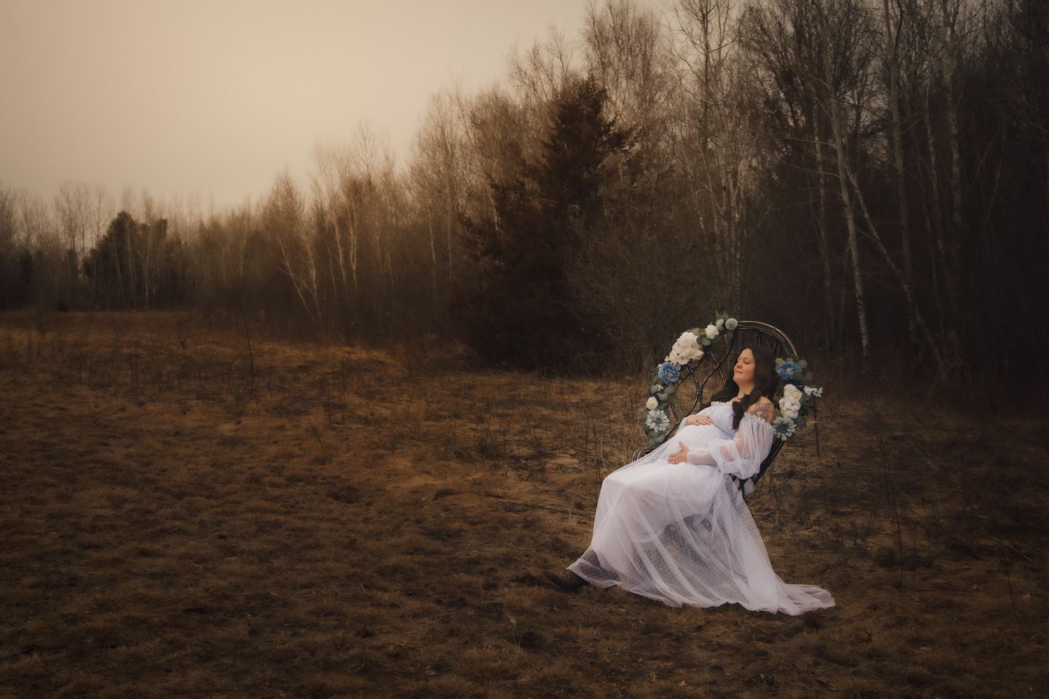 Woman in white dress sits on floral swing in misty field surrounded by bare trees.