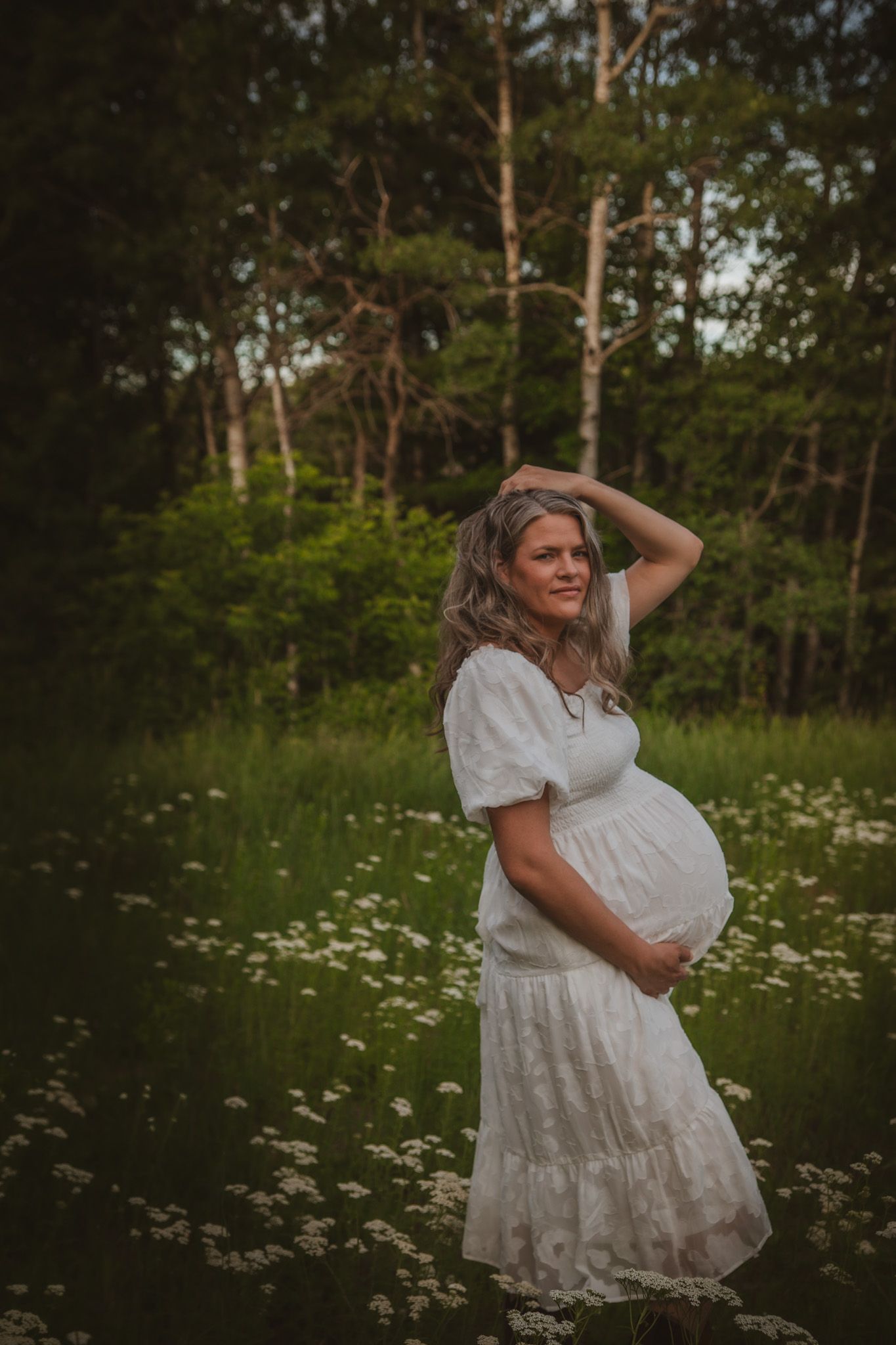Pregnant woman in white dress holding belly in a wildflower meadow with lush green forest background.