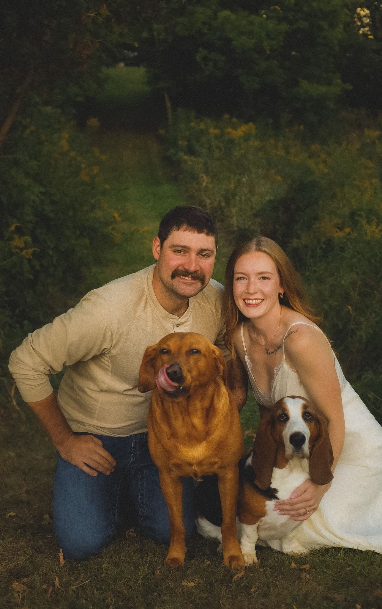 A smiling couple poses outdoors with two dogs, a golden retriever and a basset hound, surrounded by lush green foliage.