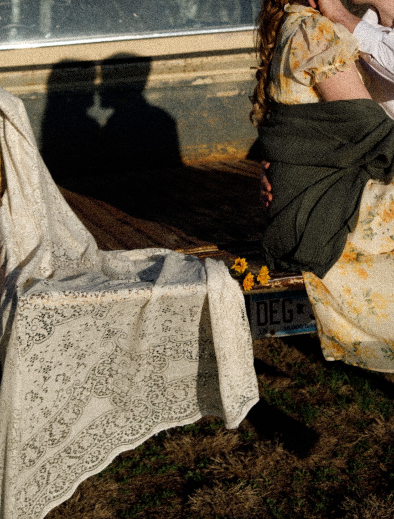 Two women in vintage floral and lace dresses stand by a car, casting romantic shadows on a sunny day.