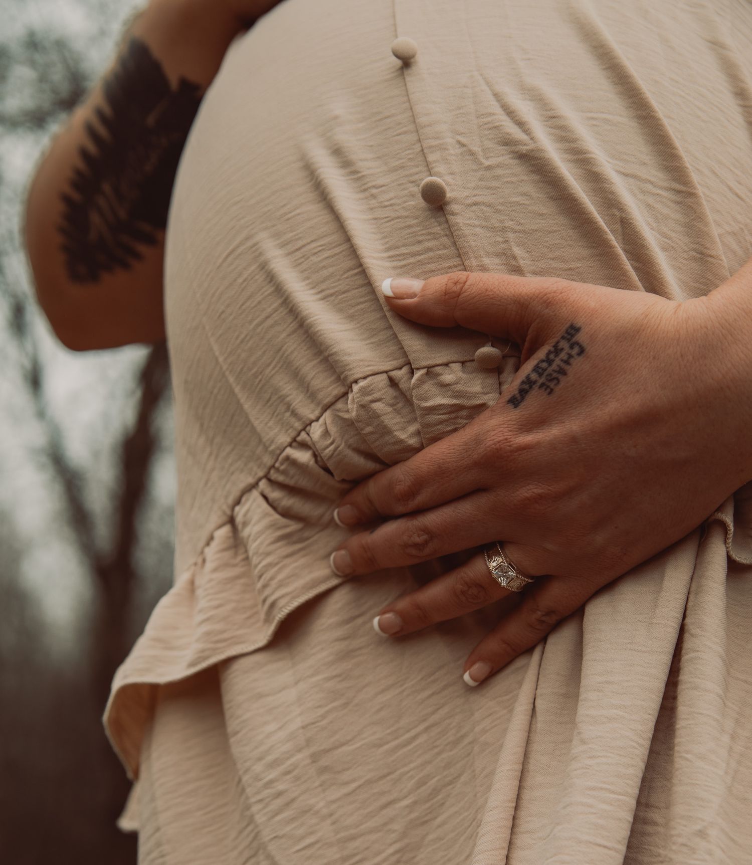 Close-up of pregnant woman in beige dress with tattoos and rings cradling baby bump outdoors.