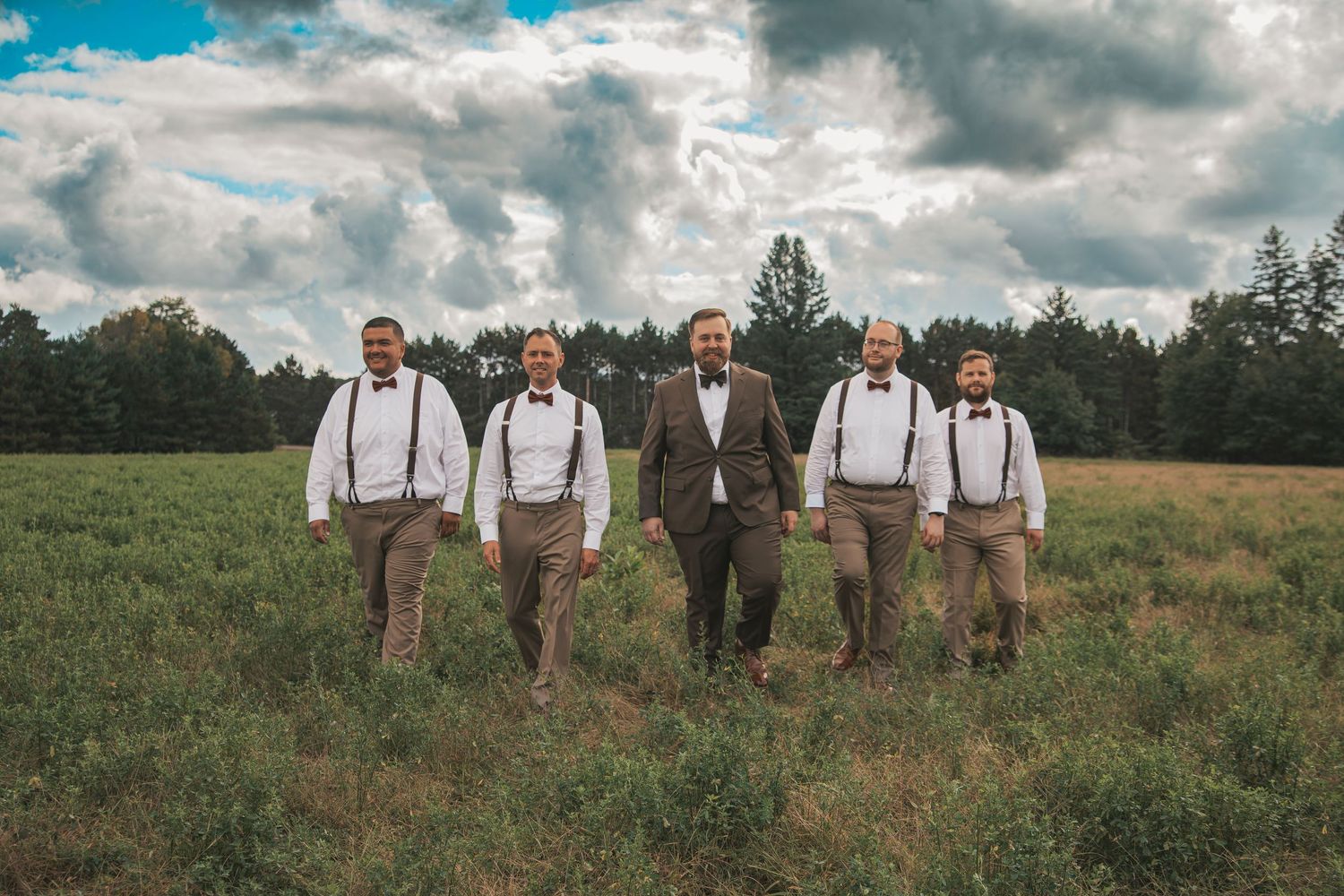 A rustic wedding party wearing suspenders and brown attire stands together in a grassy field under dramatic clouds.