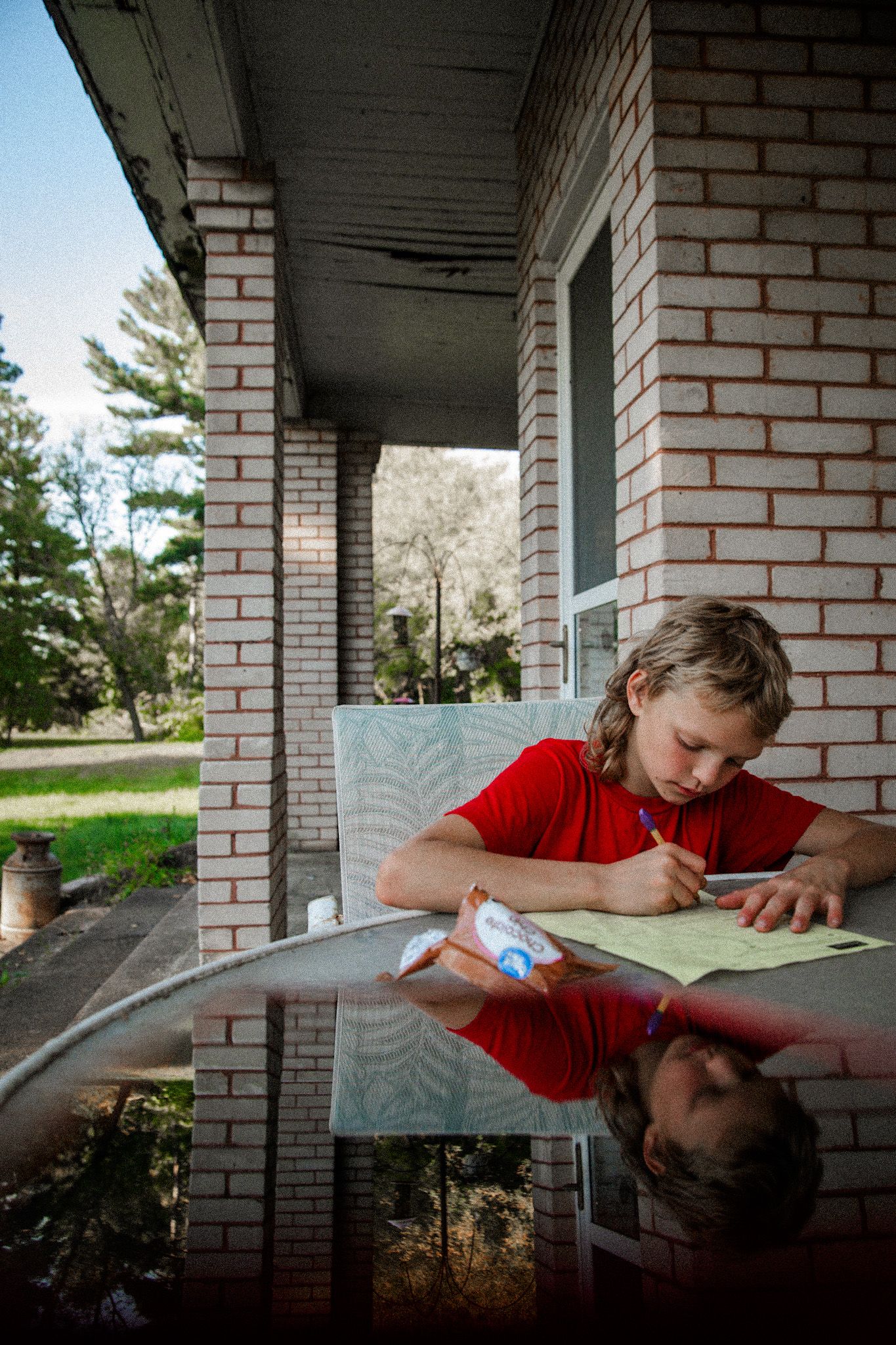 Child in red shirt doing homework at a glass table on a brick porch with reflection visible in the surface.
