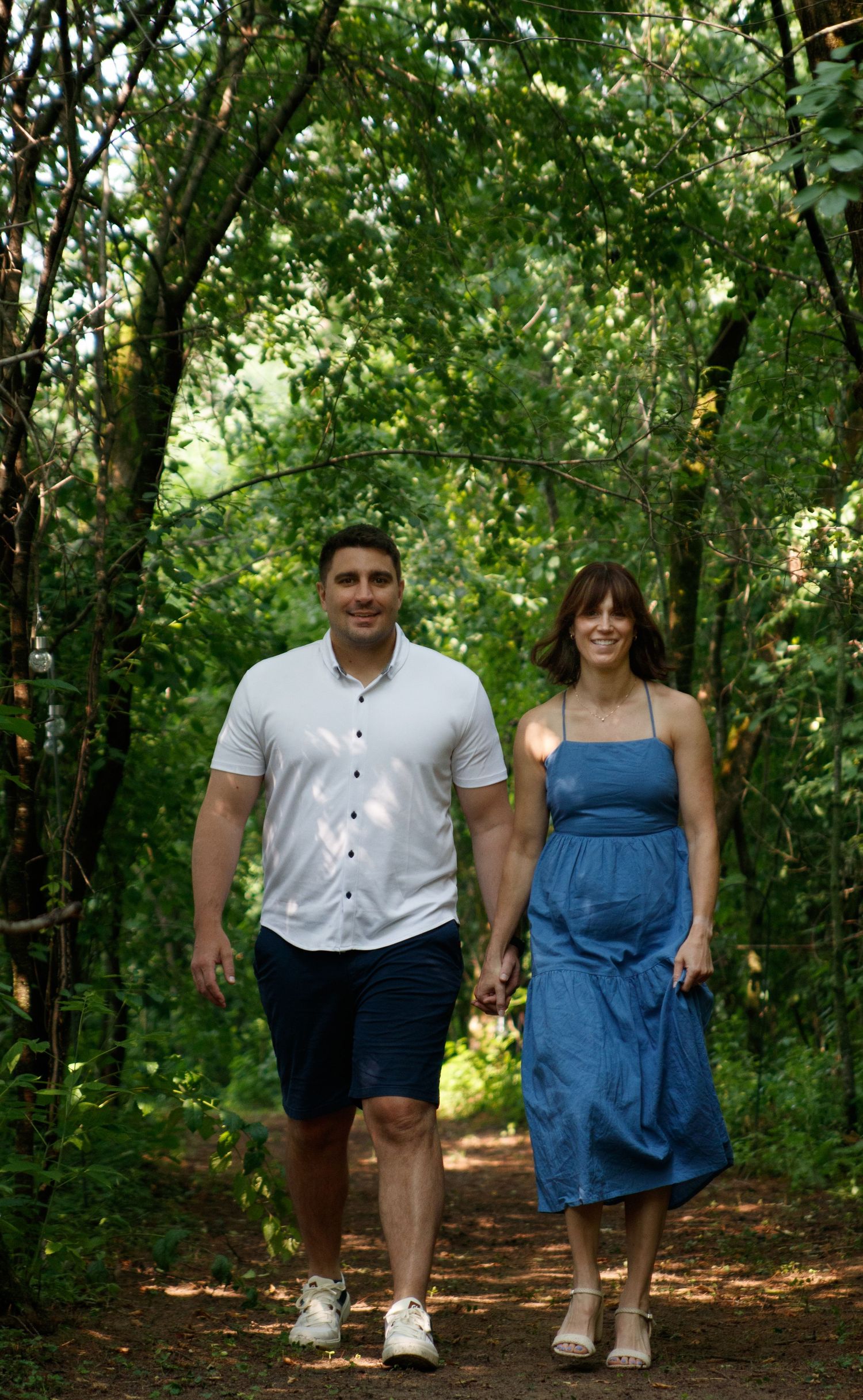 Two people walk together down a wooded trail on a sunny day, surrounded by lush green trees and foliage.