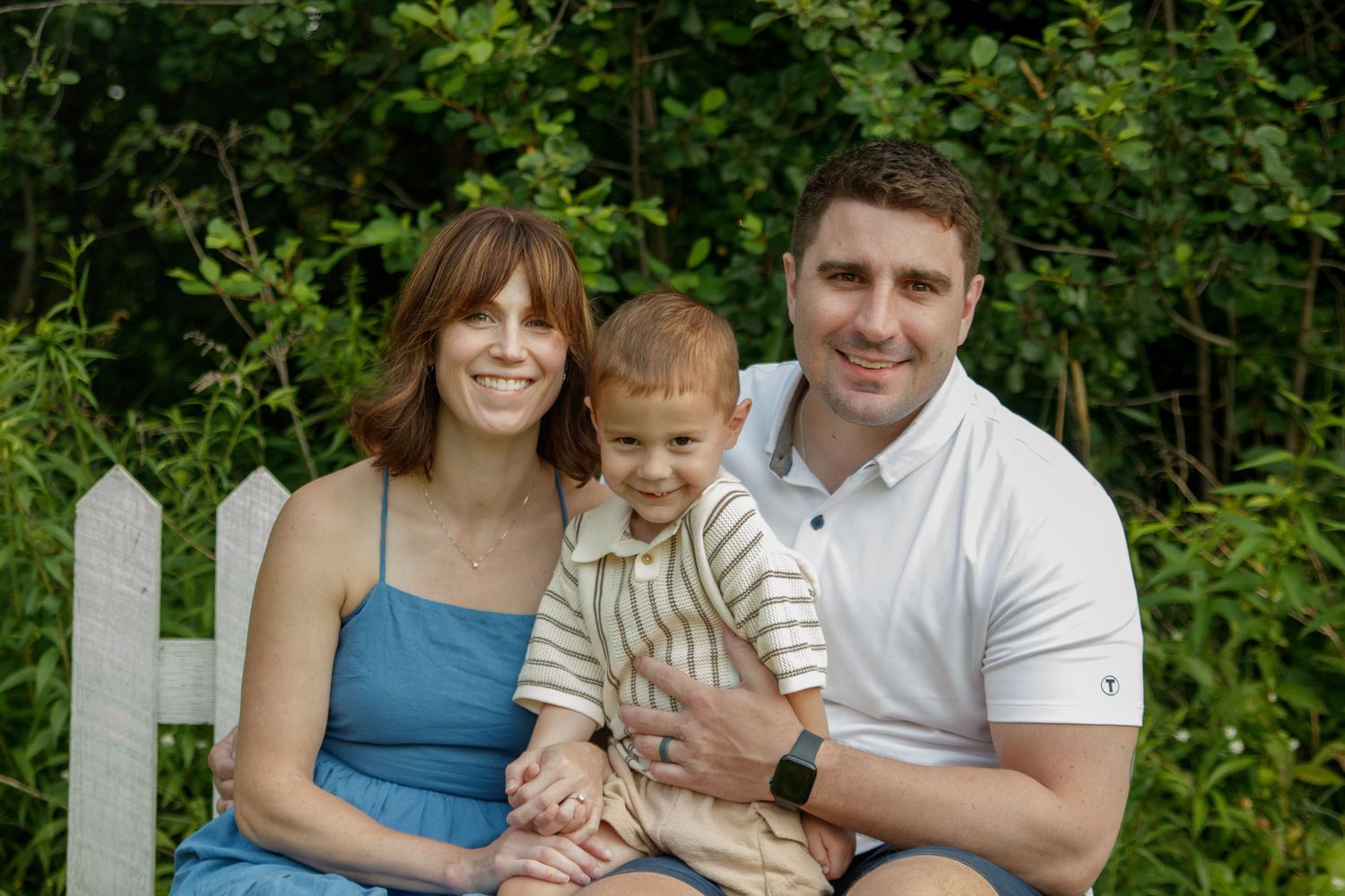 Happy family poses for a portrait together on grassy lawn near white fence.