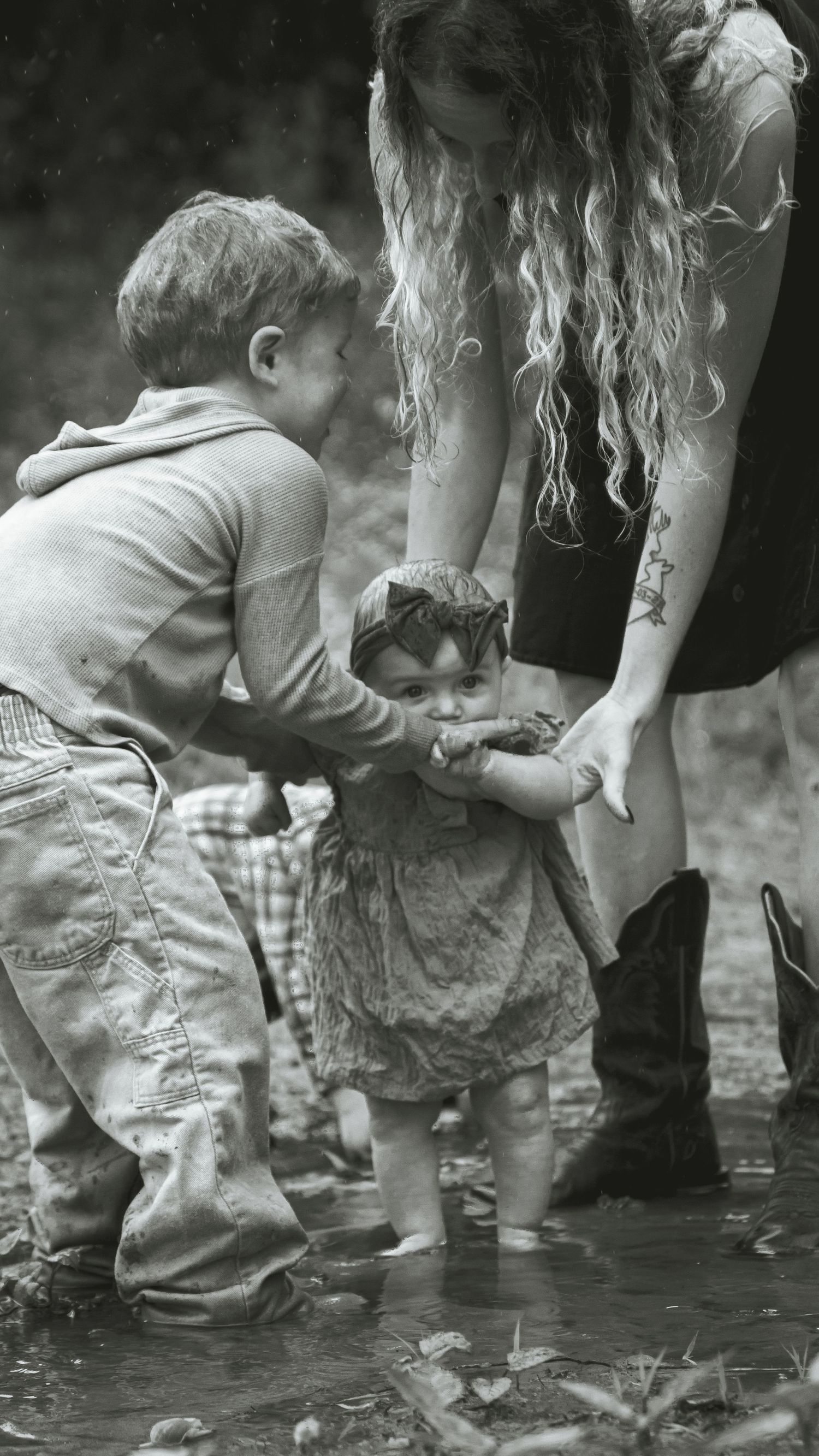 A nostalgic black and white photograph shows children and an adult splashing and playing in shallow water outdoors.