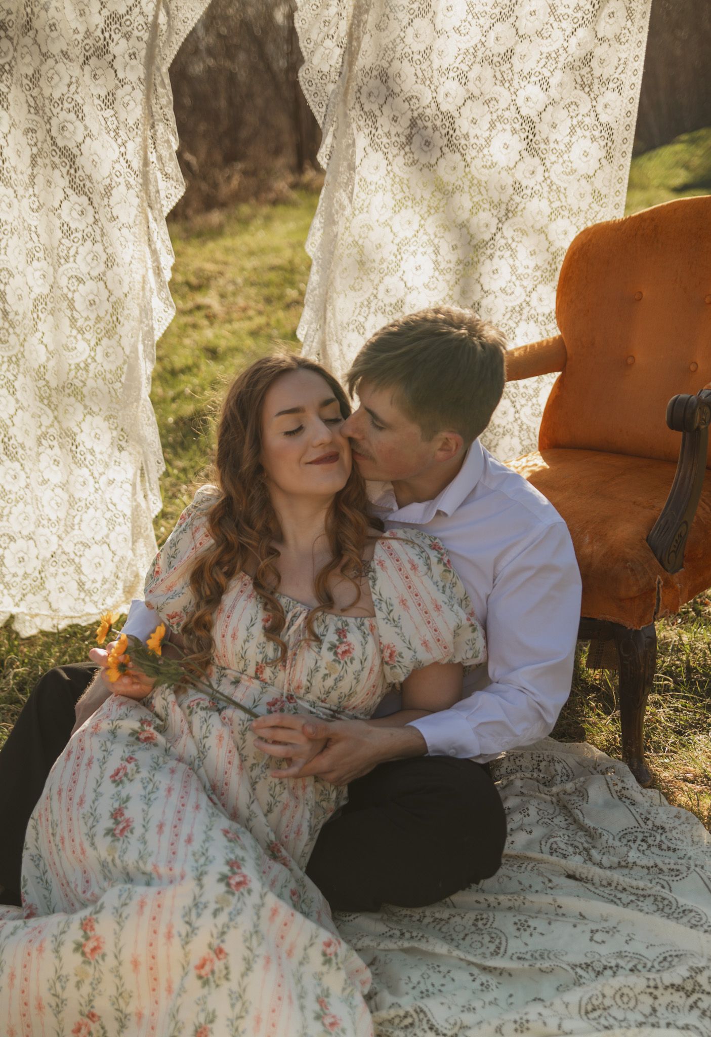 A couple sits on a blanket outdoors, man kissing woman in floral dress, lace curtains and orange chair in background.