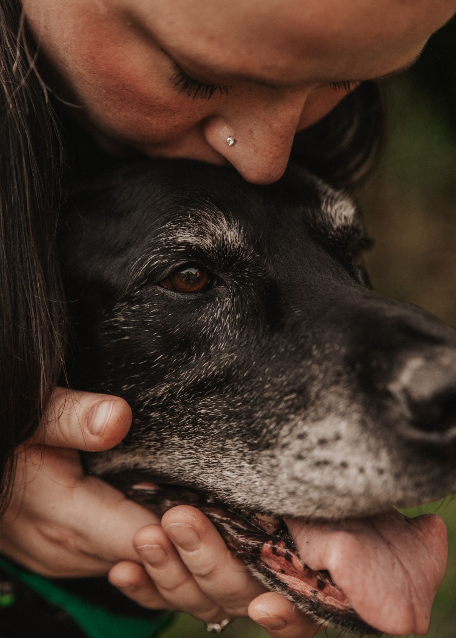 A woman with a nose piercing tenderly kisses her black dog on the head while cradling its face in her hands.