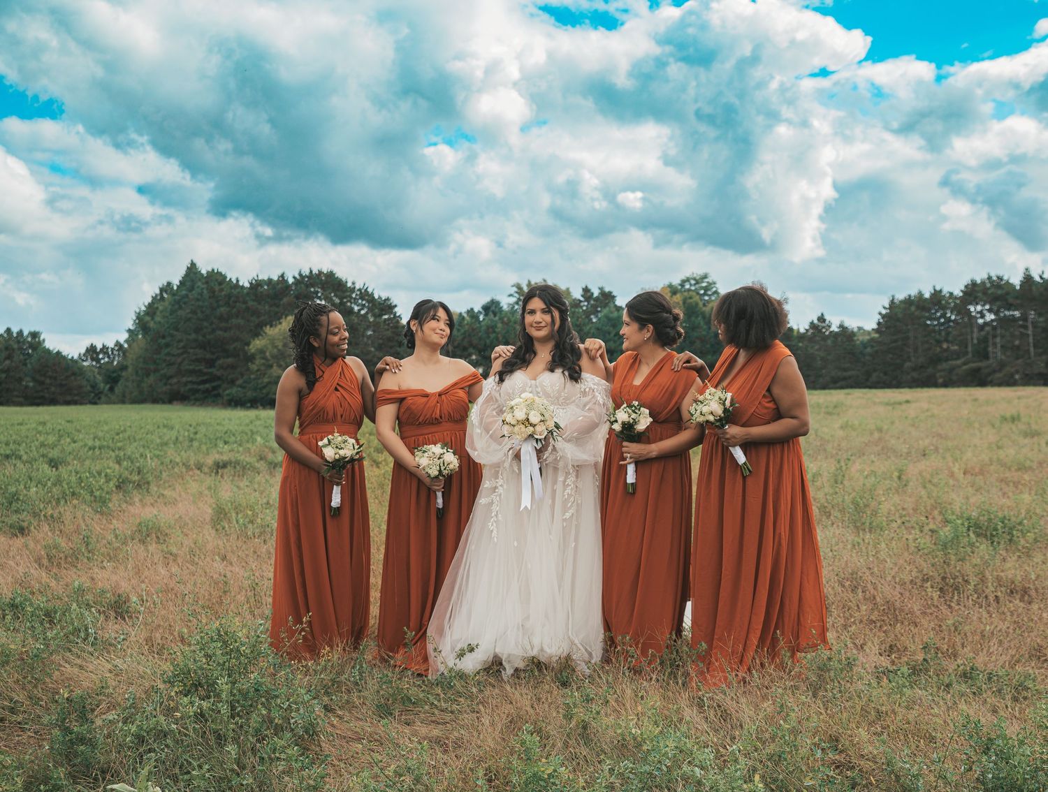 Wedding party in rust-colored bridesmaid dresses poses together in a scenic field under blue skies.