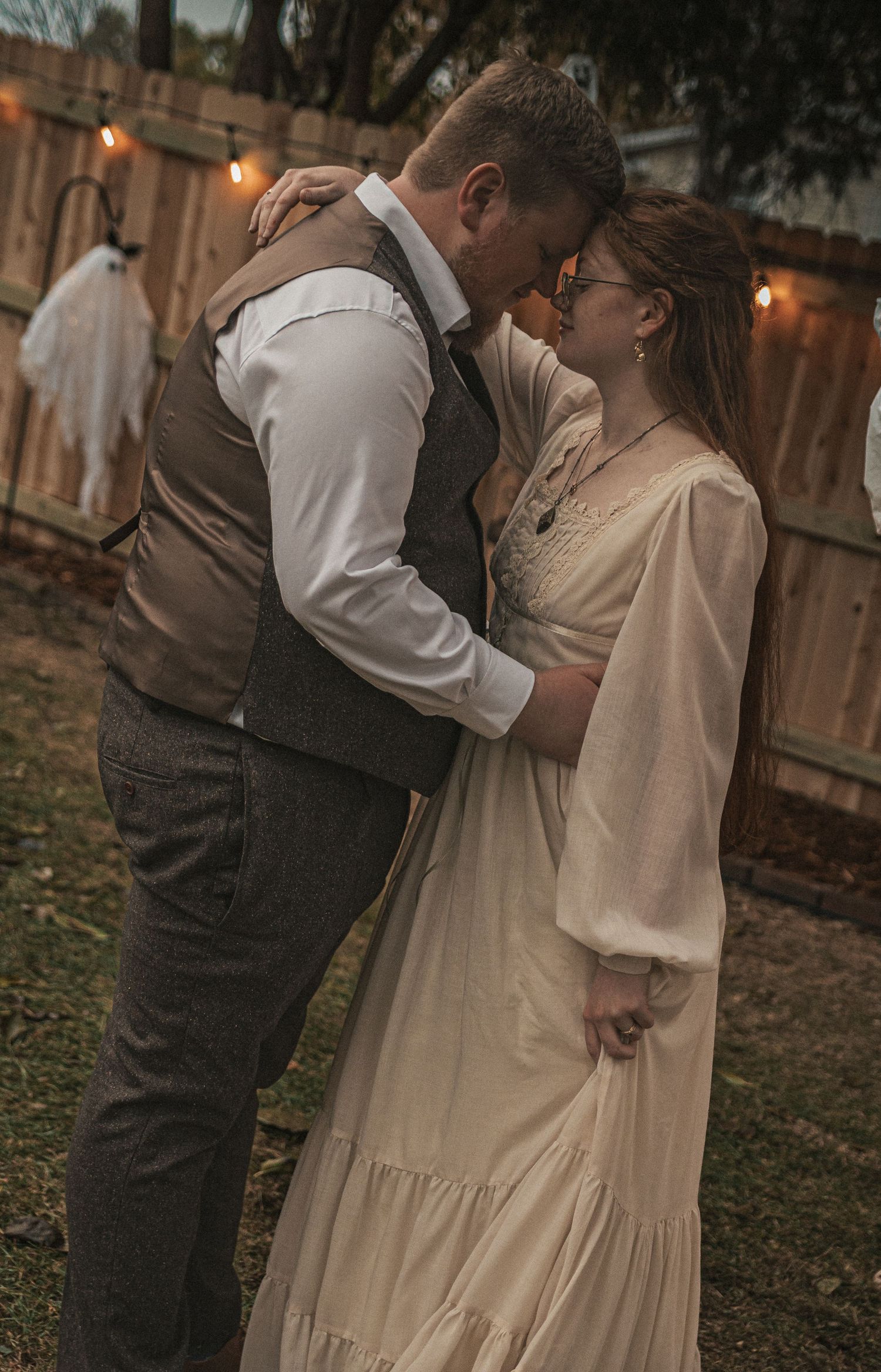 A couple shares an intimate embrace in a backyard garden at dusk with warm lighting and rustic ambiance.