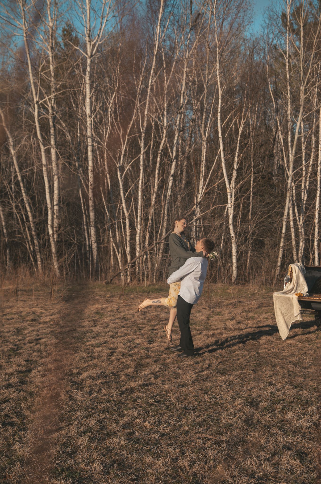 Couple embracing in a sunlit birch forest clearing during autumn, with a blanket-draped bench nearby.