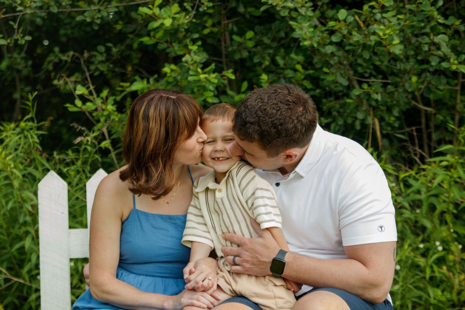 A candid family moment shared on a white picket fence surrounded by lush greenery on a summer day.