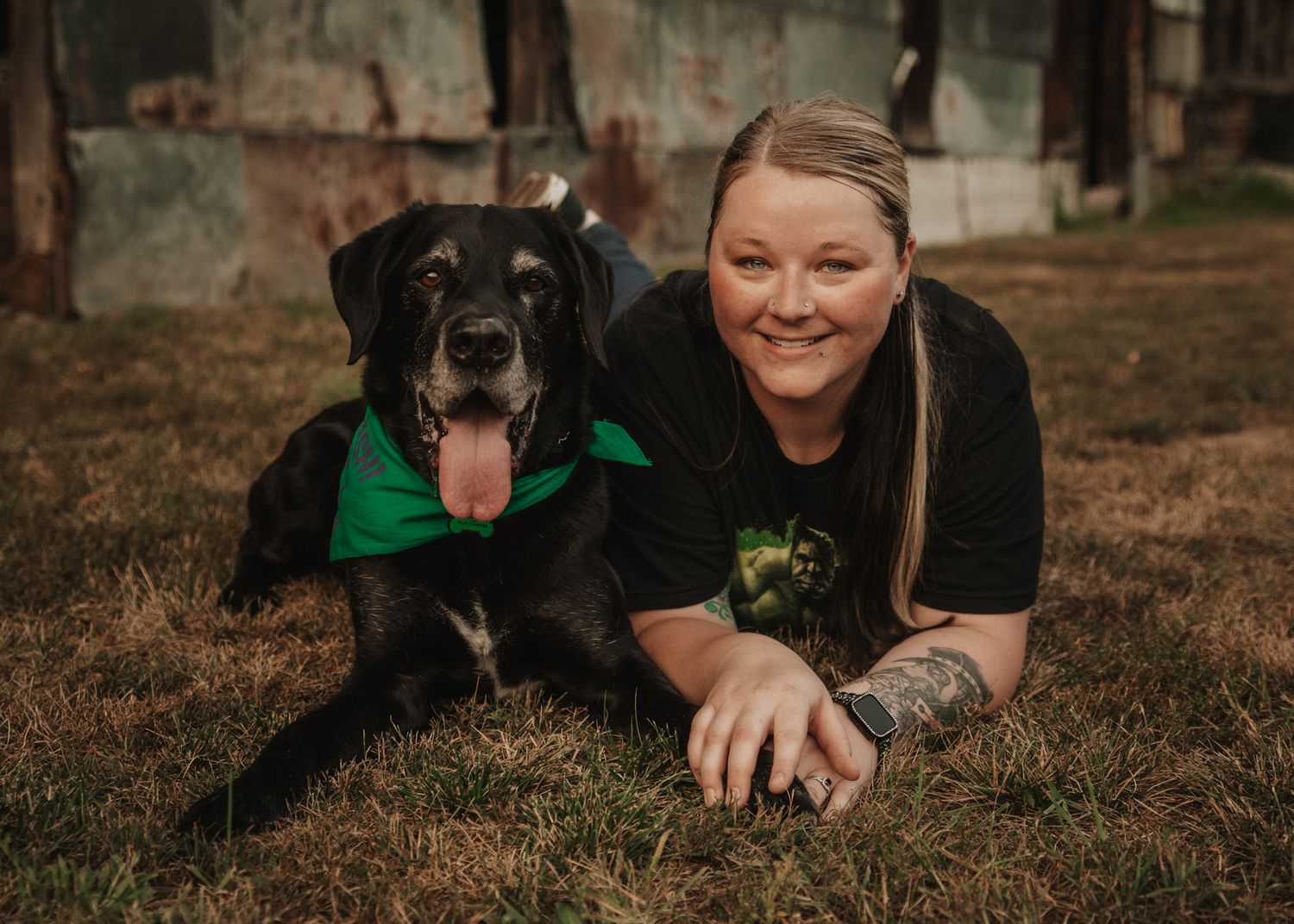Woman lying on grass smiling with black Labrador wearing green bandana outdoors.