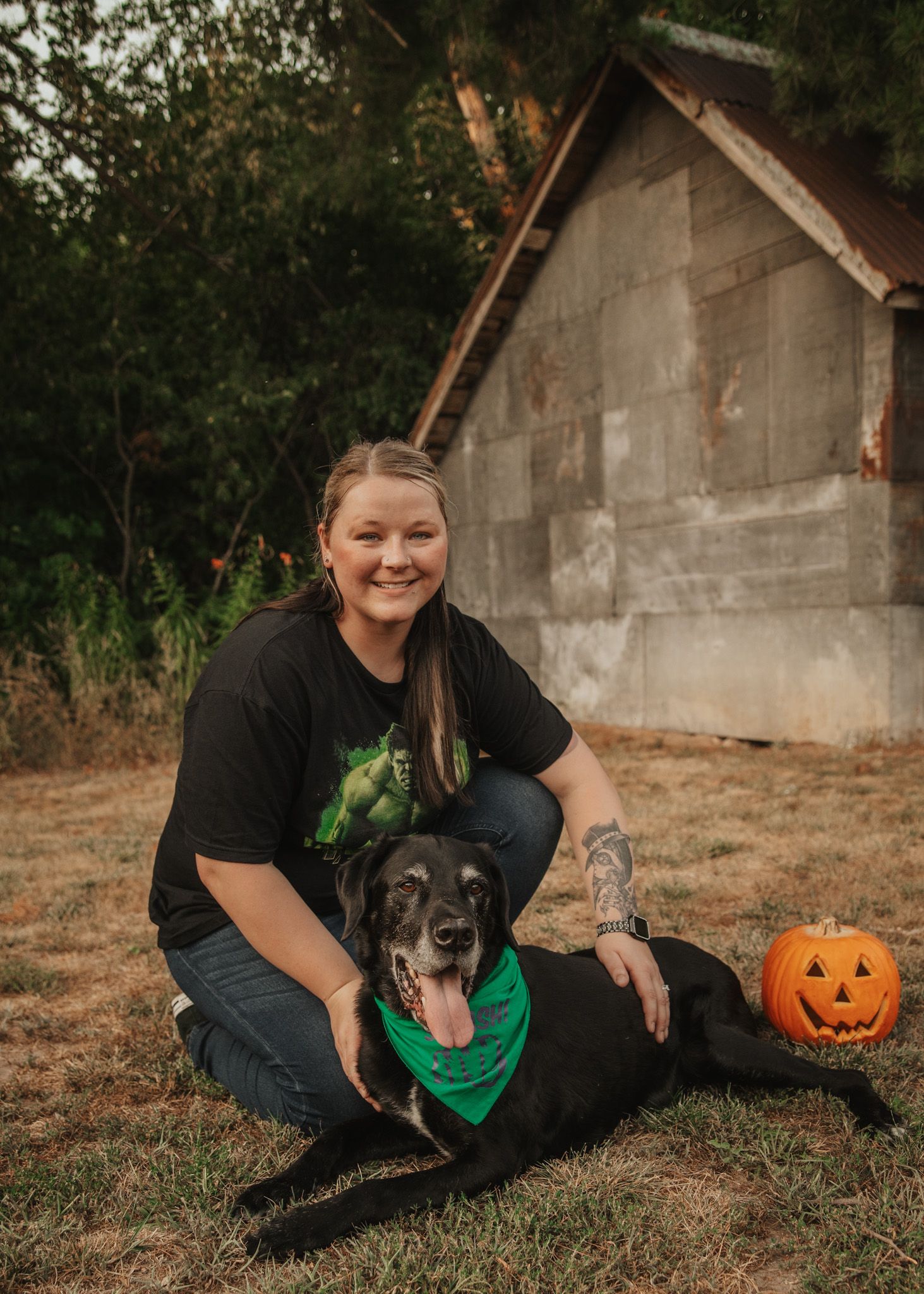 Woman sitting on grass with her black dog and a Halloween pumpkin near a rustic building.