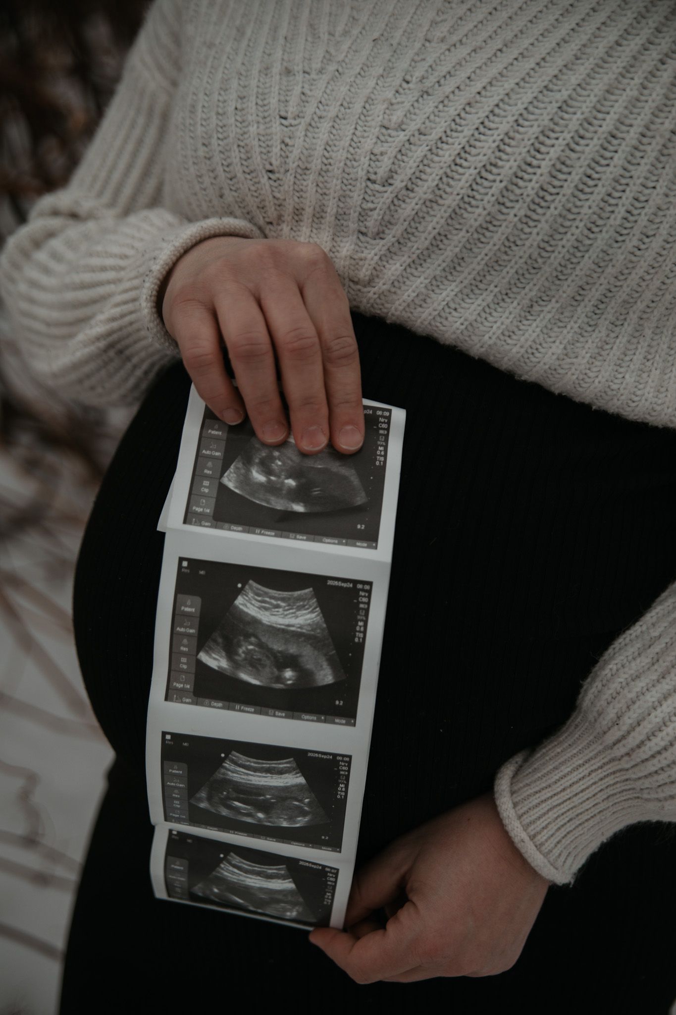 Hands holding ultrasound photos against a white knit sweater background, revealing a pregnancy announcement moment.