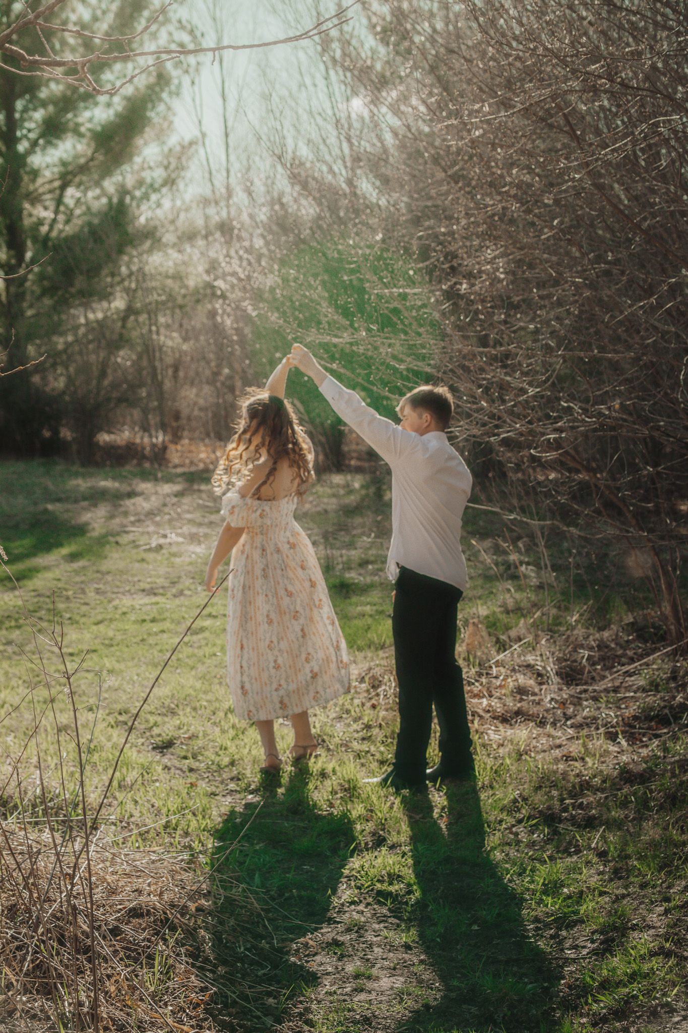 Couple dancing outdoors in sunlight, woman in floral dress twirling, man in white shirt, surrounded by trees.
