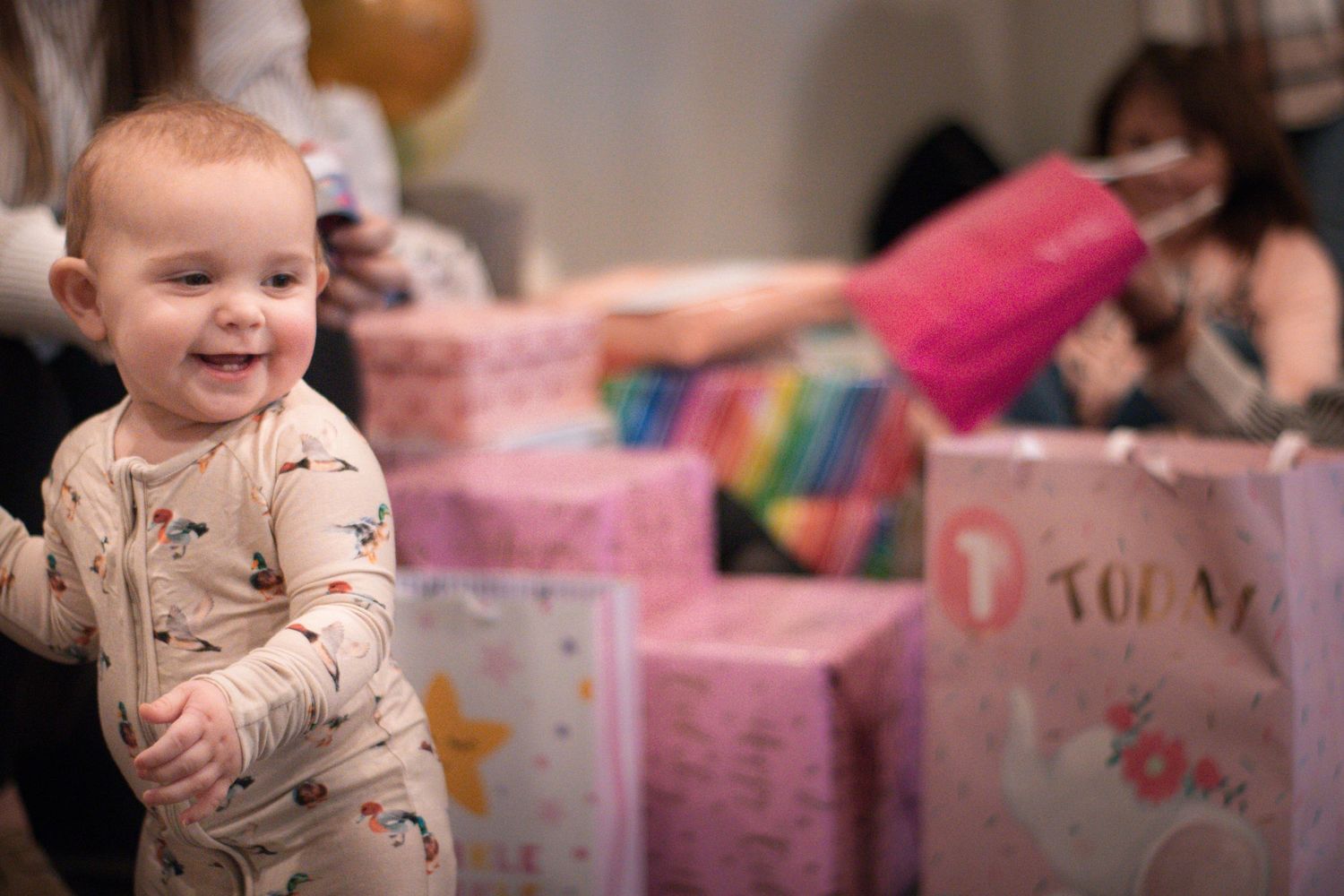 Baby in patterned pajamas smiles with pink gift boxes in background.