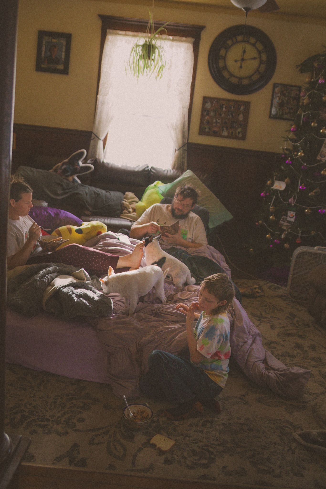 Family relaxing in cozy living room with Christmas tree, children playing with puppy on floor near bed.
