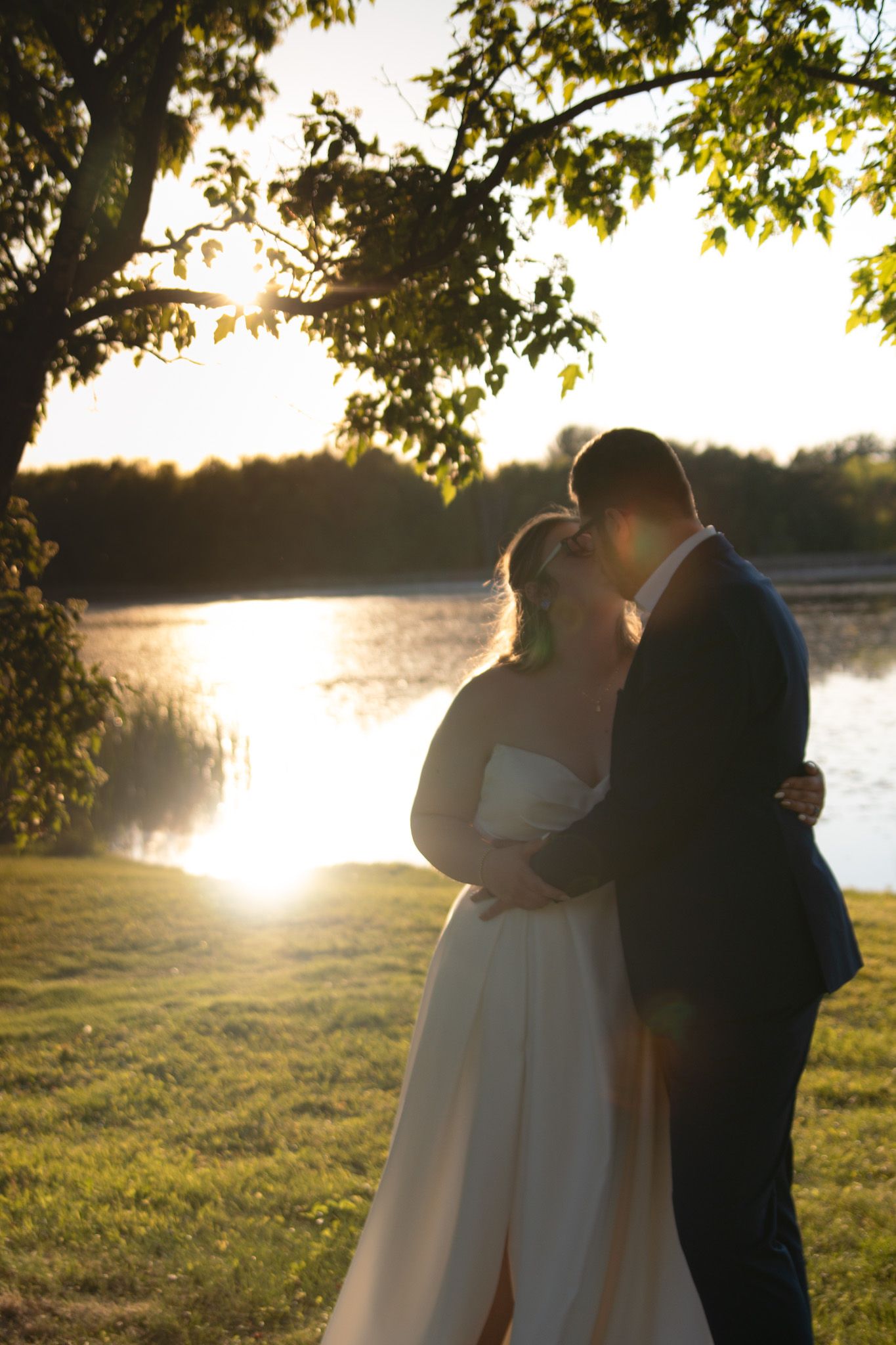 A couple embraces during sunset by a lake, with golden sunlight filtering through tree branches in a romantic outdoor setting.