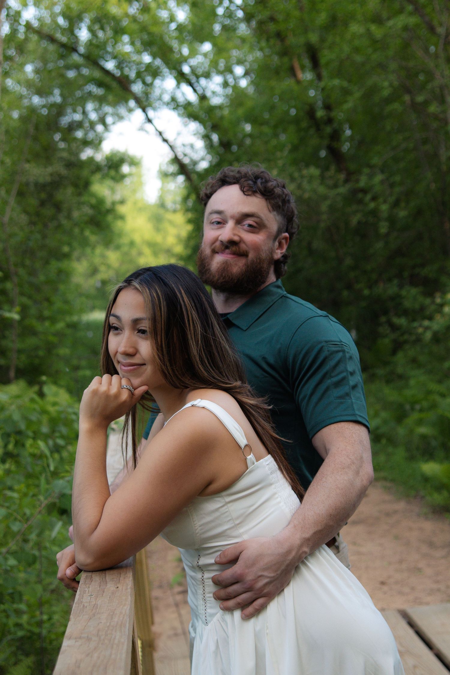 A couple shares a tender moment on a wooden bridge surrounded by lush green trees in a natural setting.