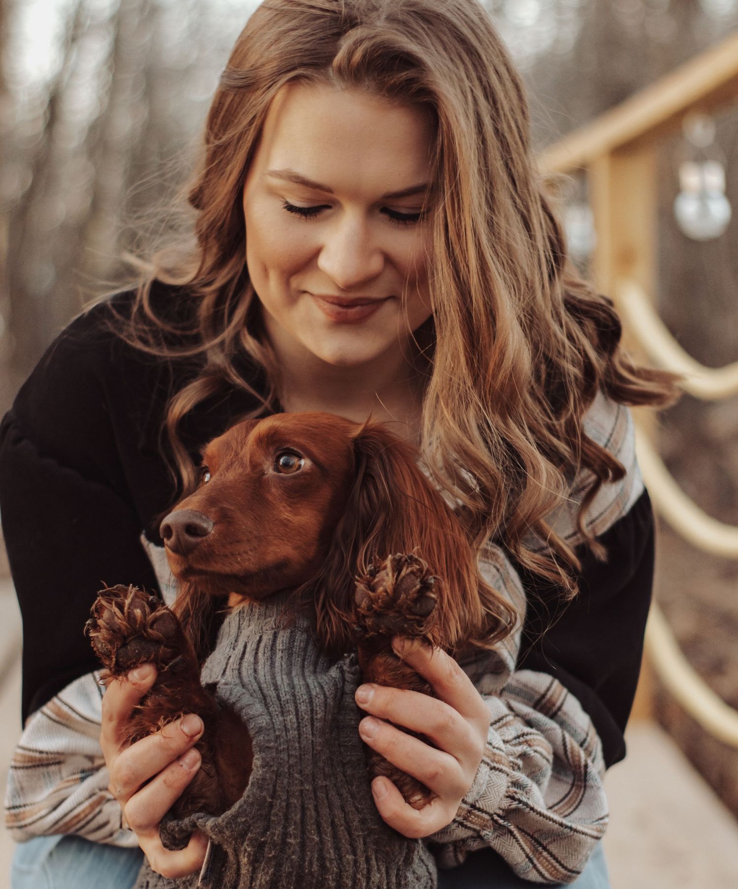 A person in a black sweater smiles warmly while cuddling a small brown dachshund puppy wearing a gray knit sweater.