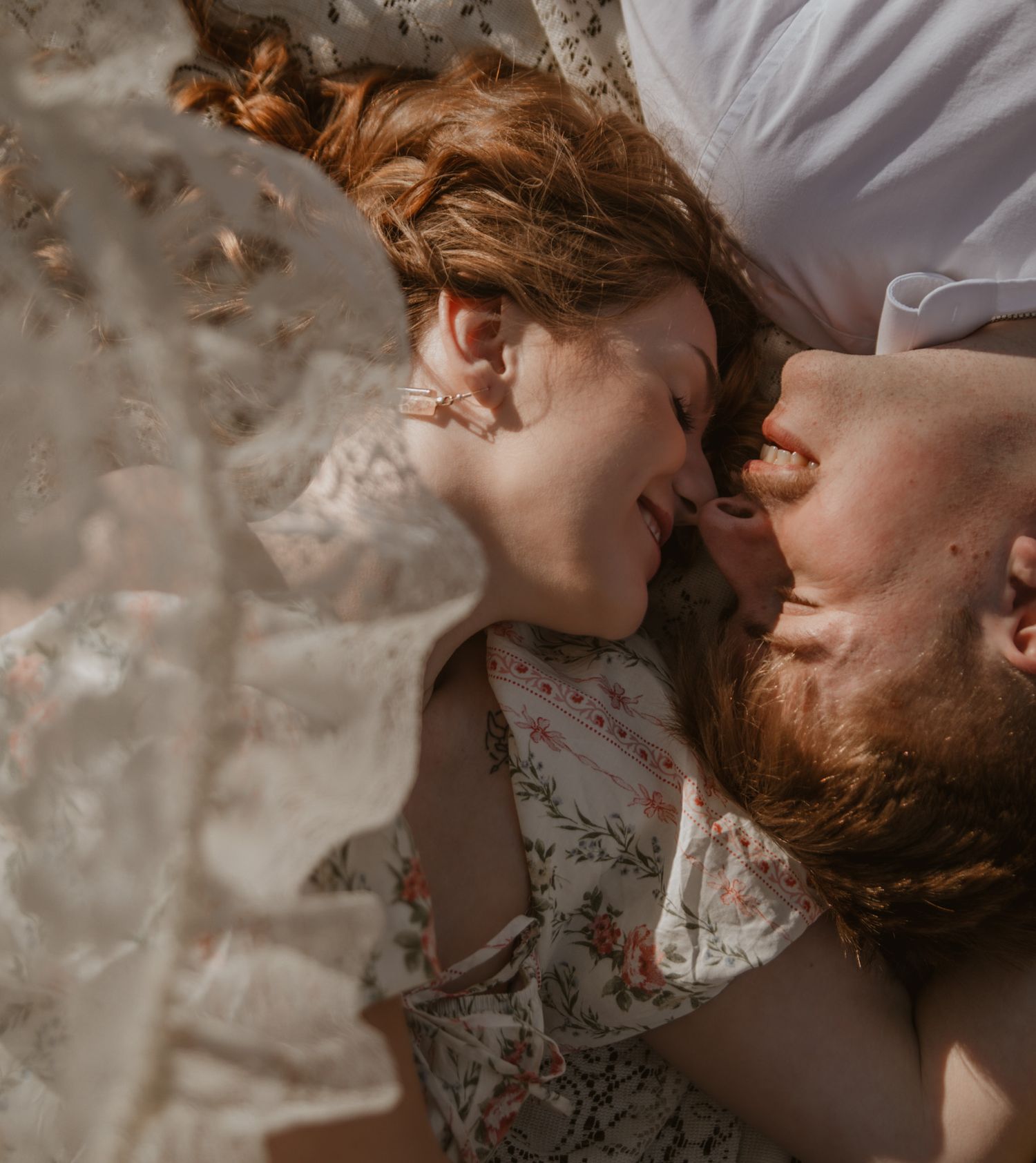 Romantic couple lying together nose-to-nose, smiling intimately, woman in floral dress with red hair.
