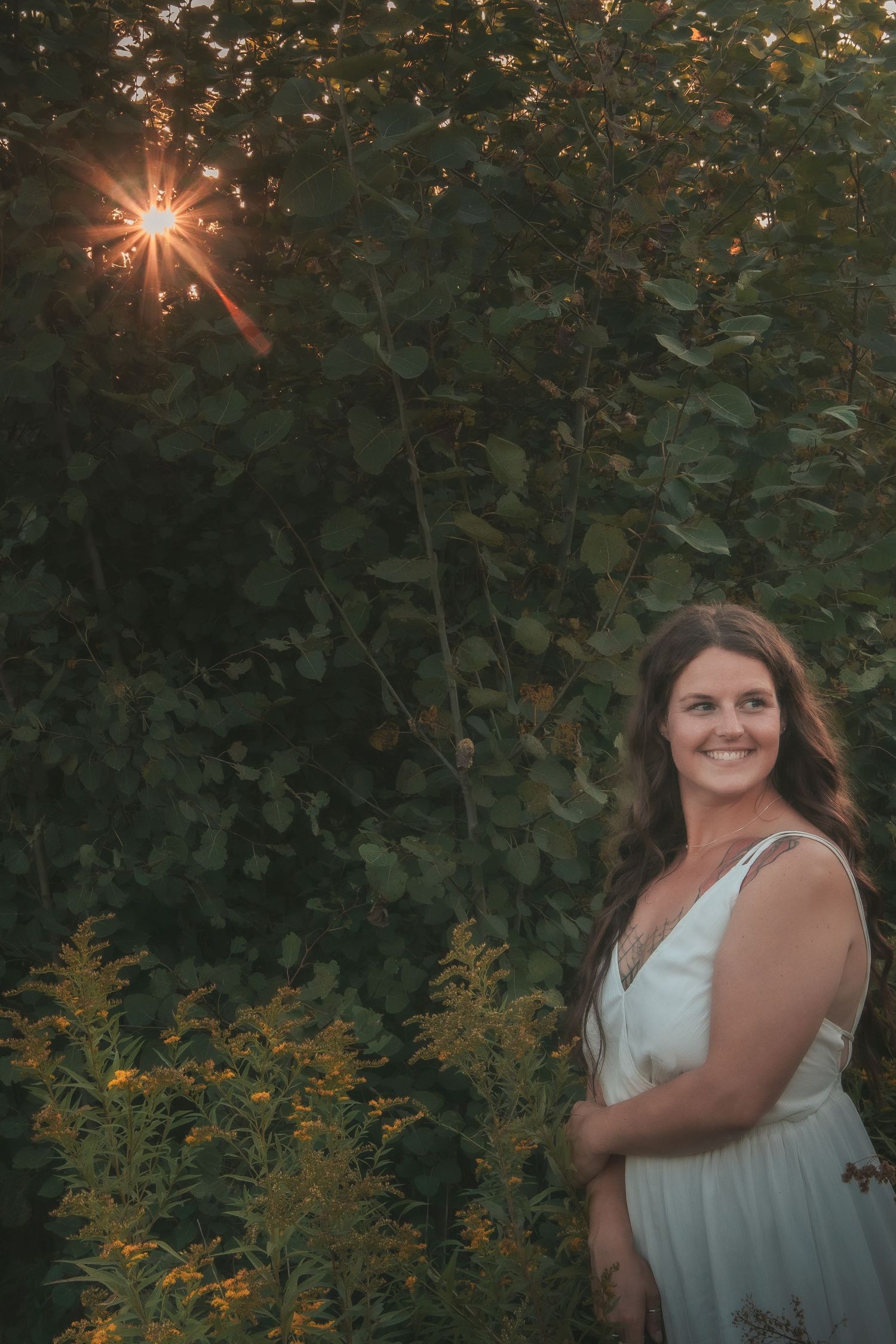 A white dress stands out against lush greenery at sunset with a starburst effect through the leaves in a garden setting.