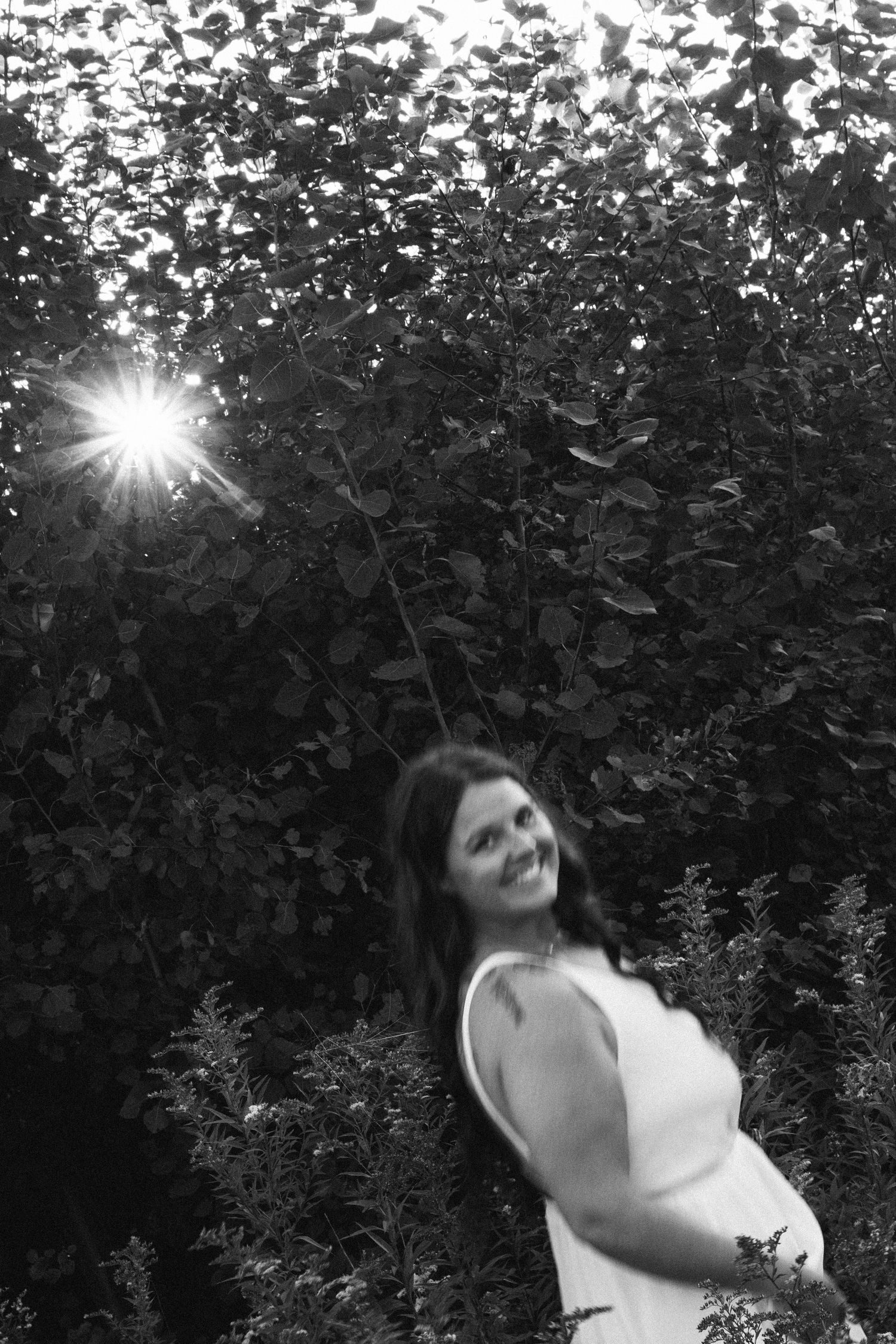 Artistic black and white nature photo of a silhouetted figure among lush foliage with dramatic sunburst shining through trees.