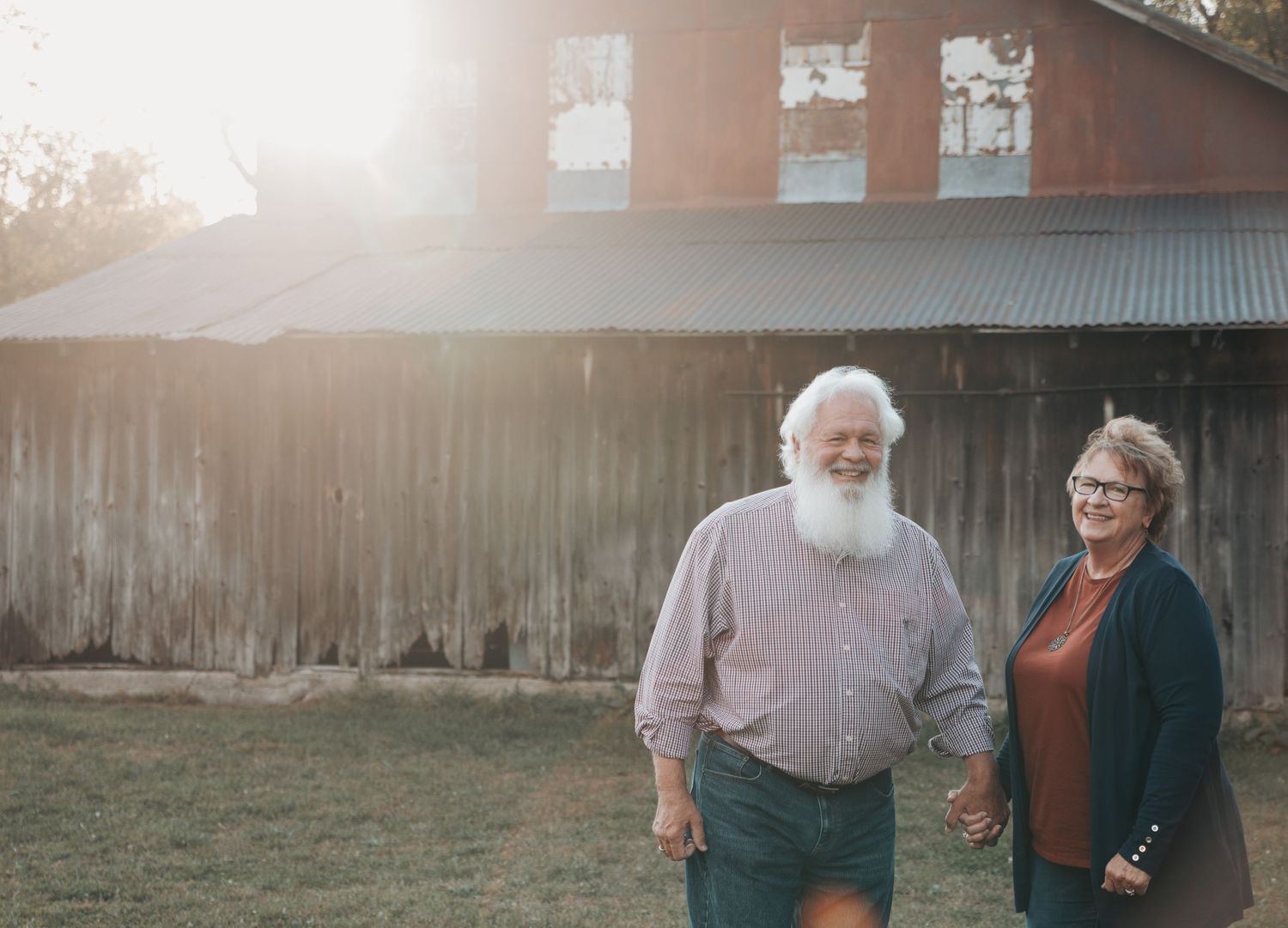 Two people walking together on a path through a misty field at sunset with a wooden fence in the background.