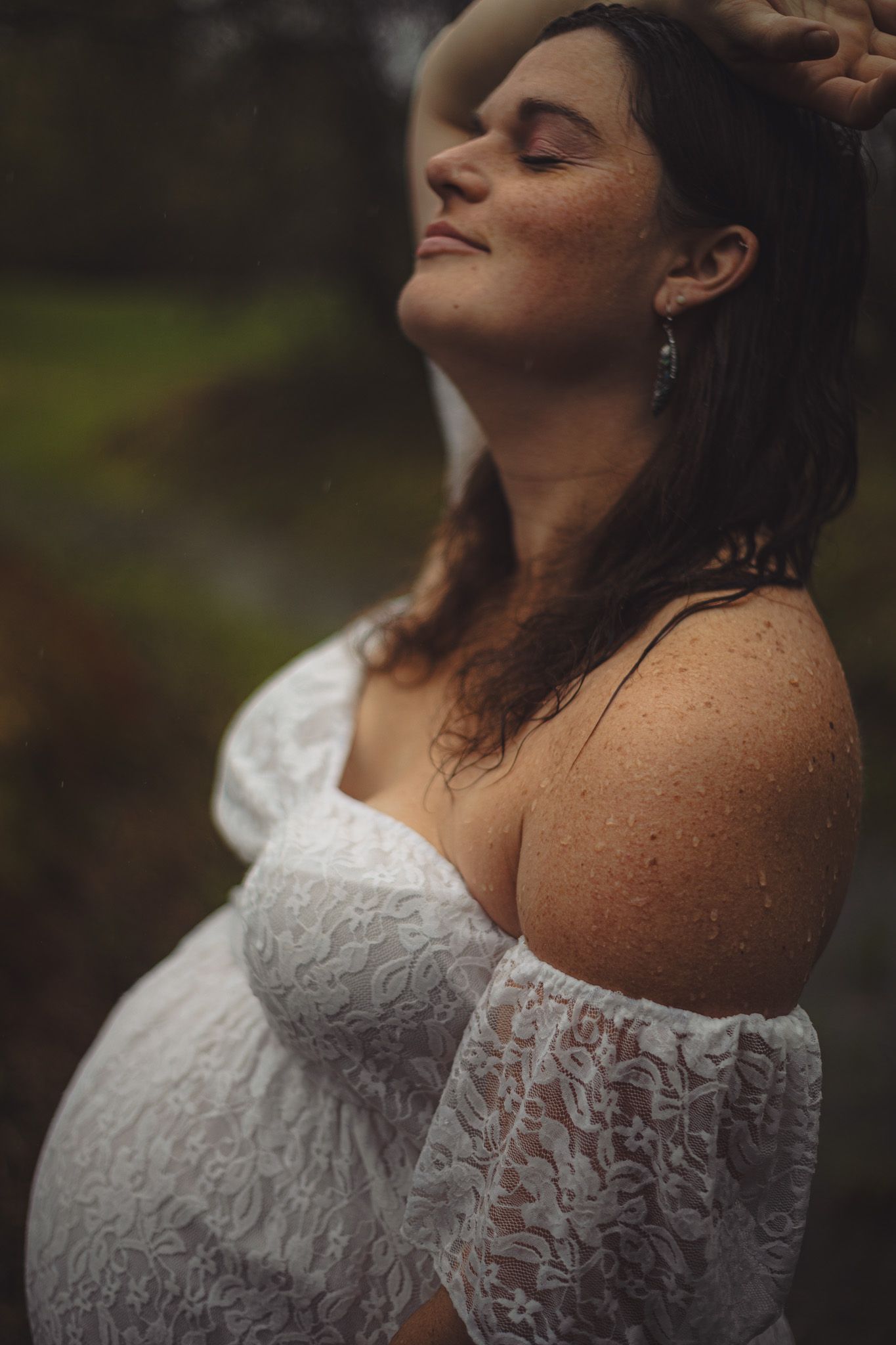 Serene pregnant woman in white lace dress with eyes closed, rain drops on skin.