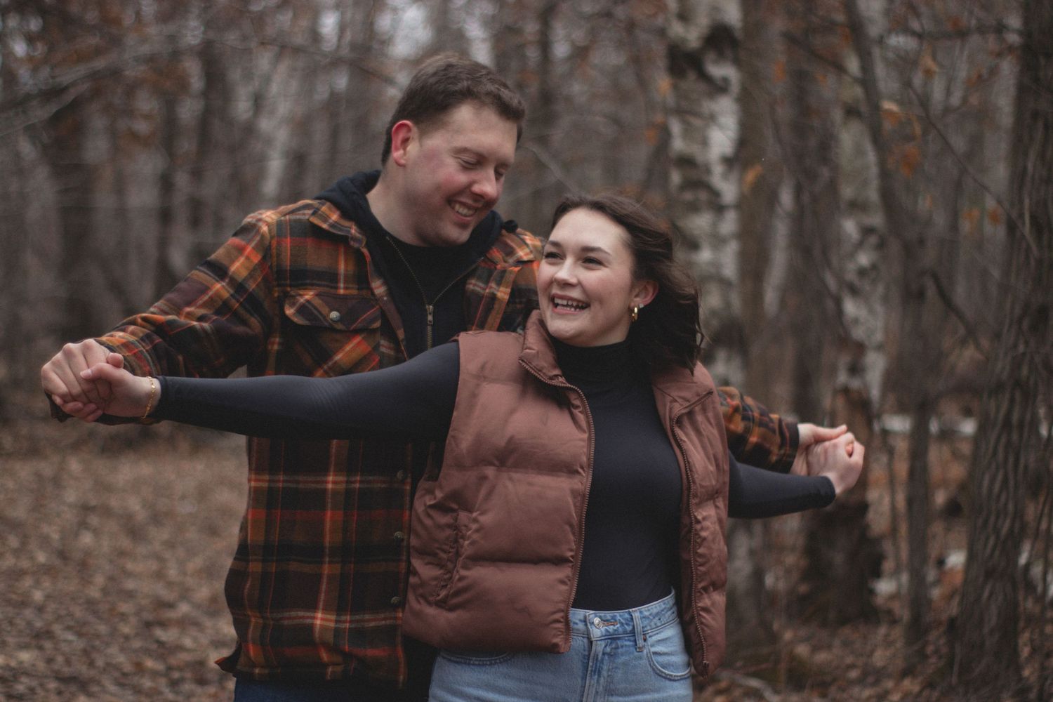 A joyful couple shares a candid laugh while walking through an autumn forest with bare trees.