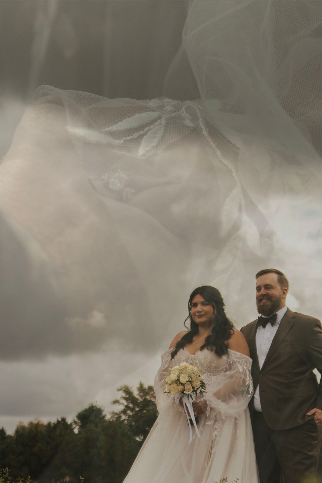 A wedding party stands outdoors against a dramatic cloudy sky in a panoramic shot.