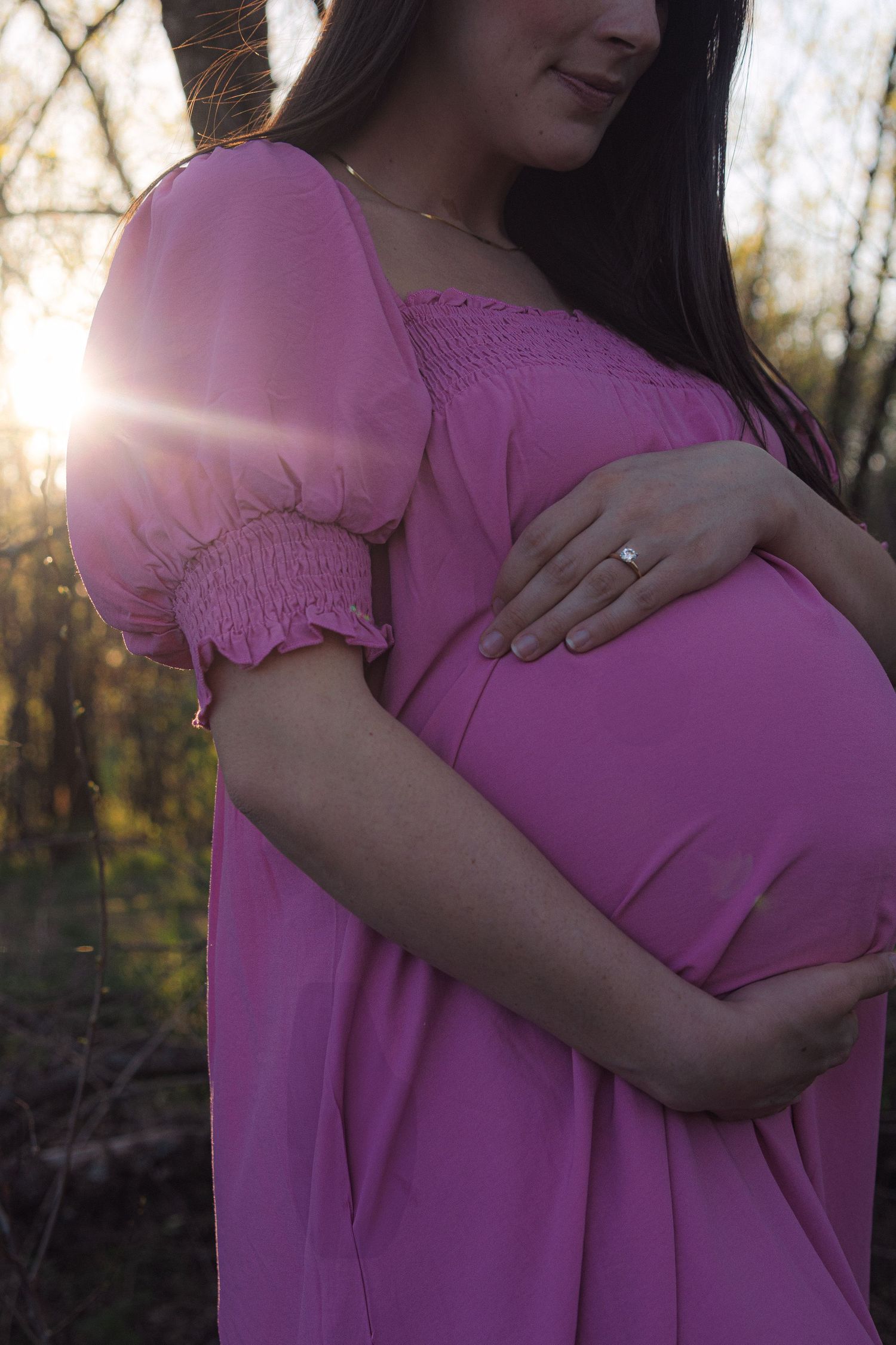 A person wearing a flowing purple dress cradles their pregnant belly in dappled sunlight among trees.