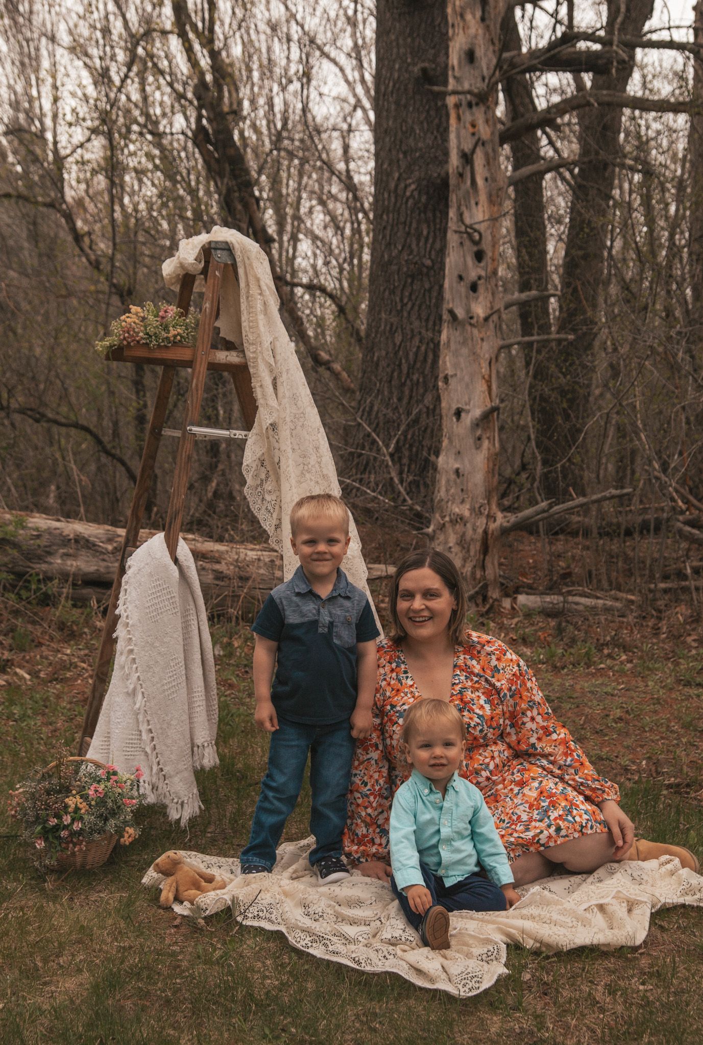 A mother in a floral dress poses outdoors with two young boys on a lace blanket near a rustic ladder in a wooded setting.