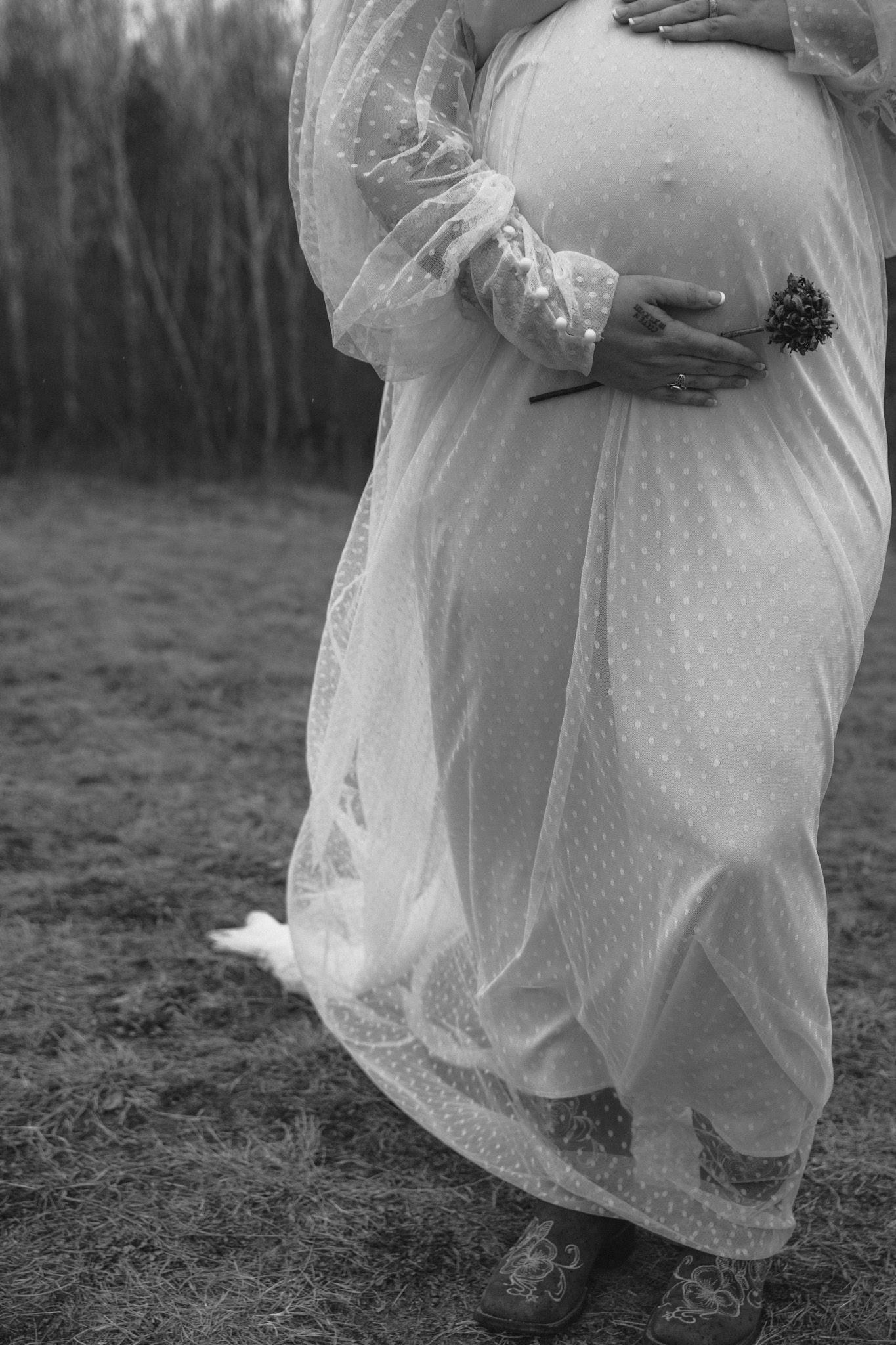 Black and white close-up of pregnant woman in sheer white polka dot gown cradling baby bump outdoors.
