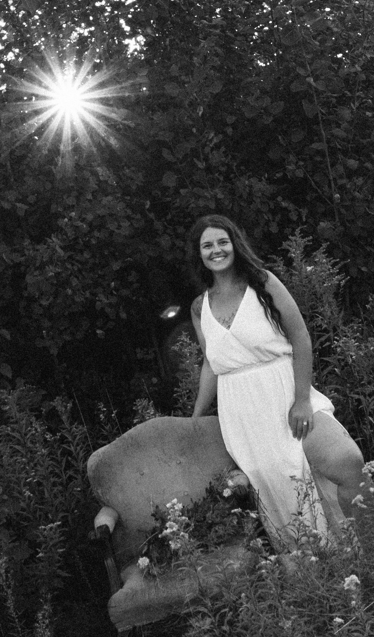 A person in a white dress poses outdoors surrounded by sunlit foliage in a black and white photograph.