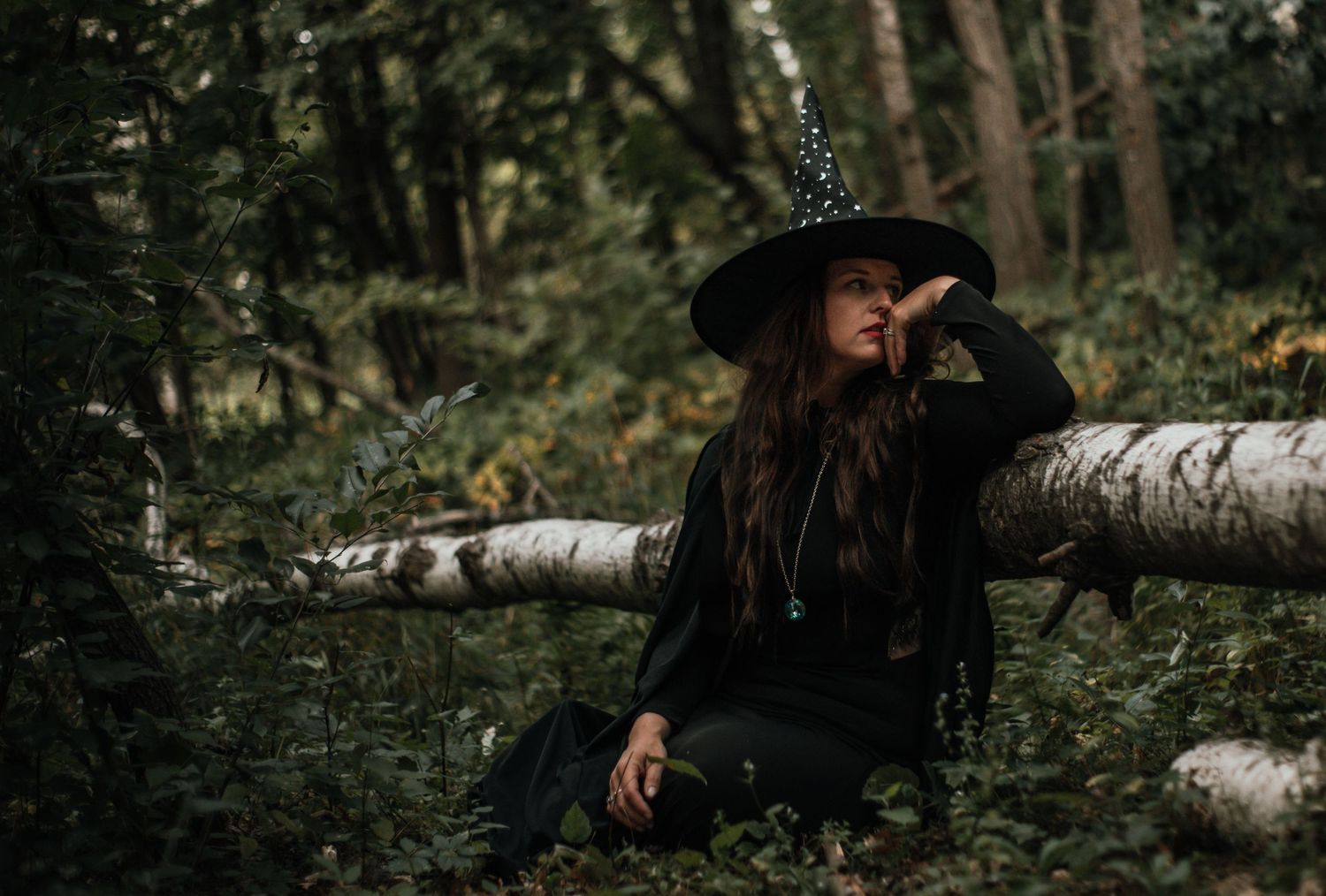 A figure in a dark hat lies contemplatively on a fallen birch tree trunk in a lush green forest setting.