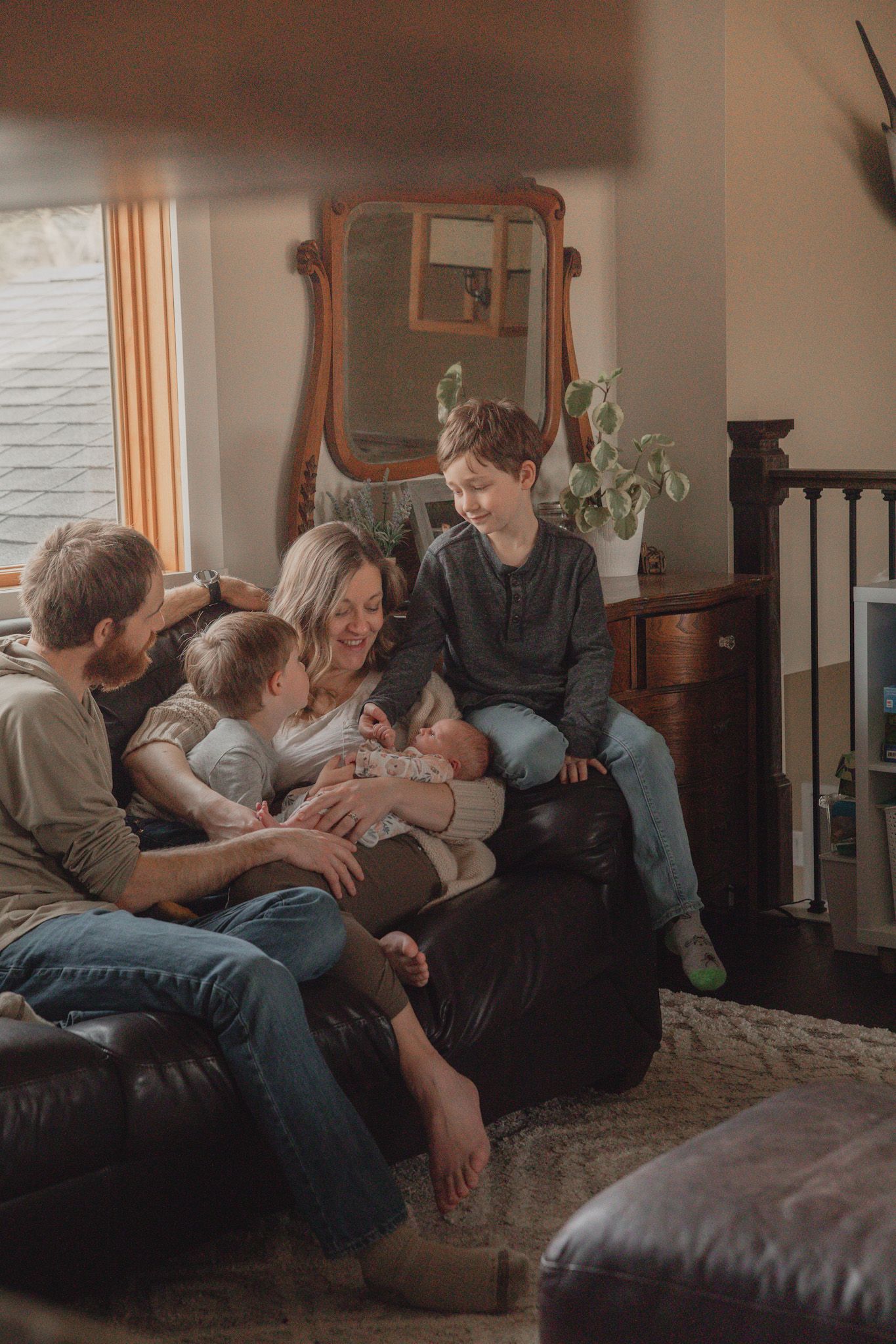 Family of five gathered on a leather sofa in a cozy living room, parents and children admiring a newborn baby.