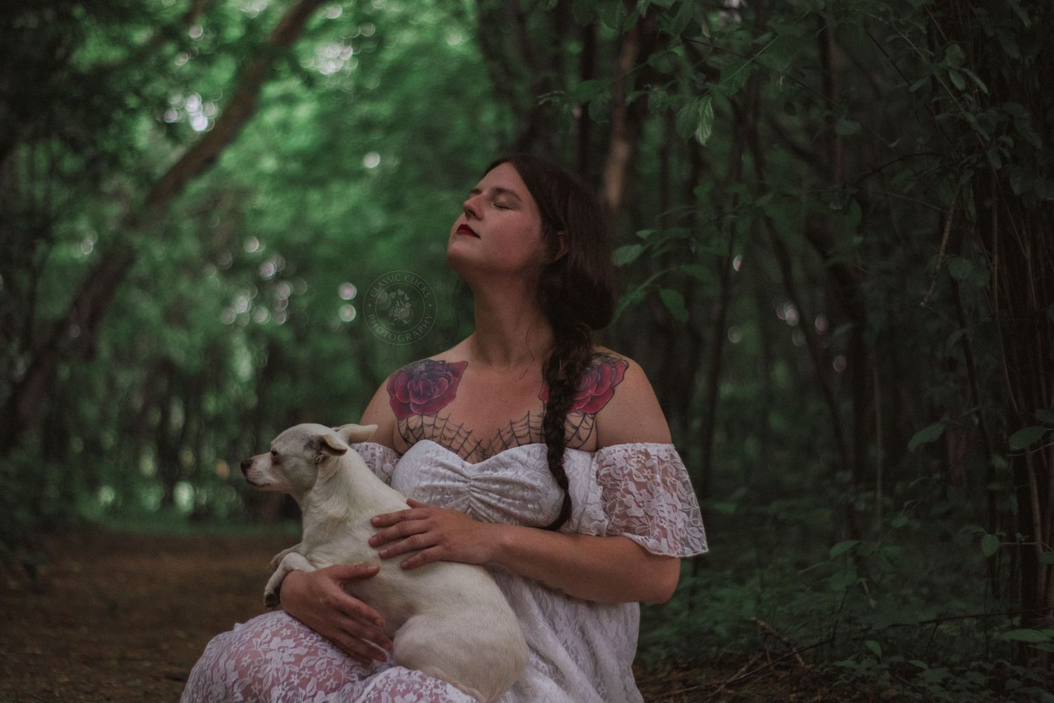 A person in a floral dress holds a white cat while standing in a lush green forest setting.