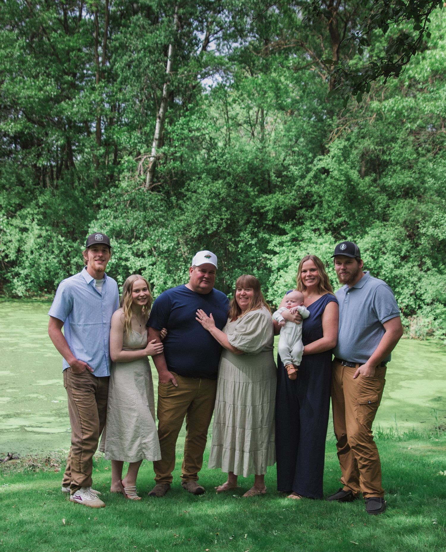 A group poses together outdoors in formal attire on a sunny day with green trees and grass in the background.