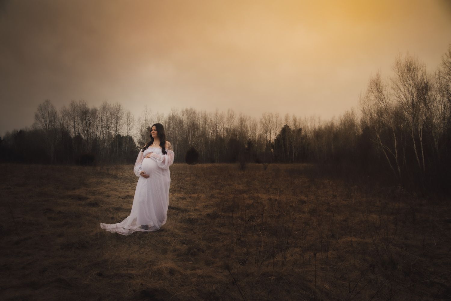 Pregnant woman in white lace maternity gown standing in golden field at sunset.