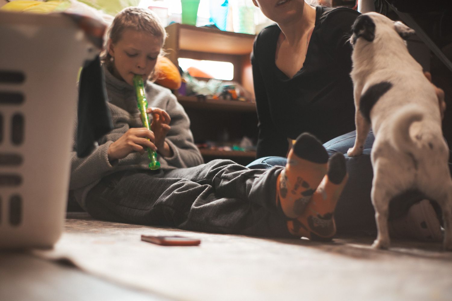 Children and a dog play on the floor together indoors while a woman watches nearby in a cozy home setting.