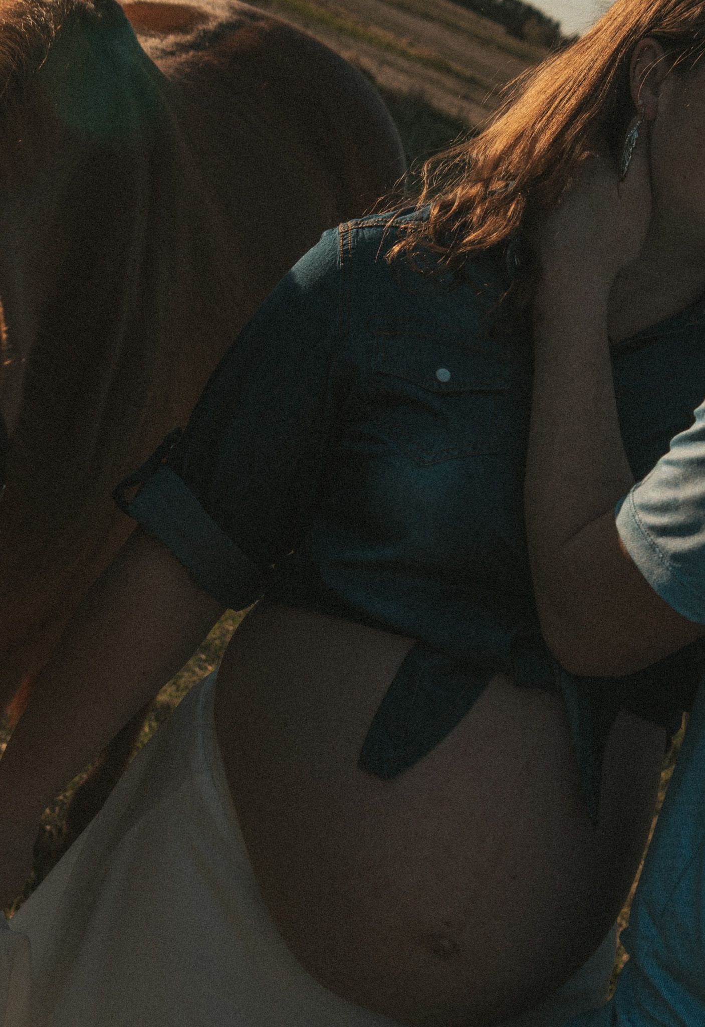 Close-up of a pregnant woman's bare baby bump, wearing a denim shirt outdoors in warm golden light.