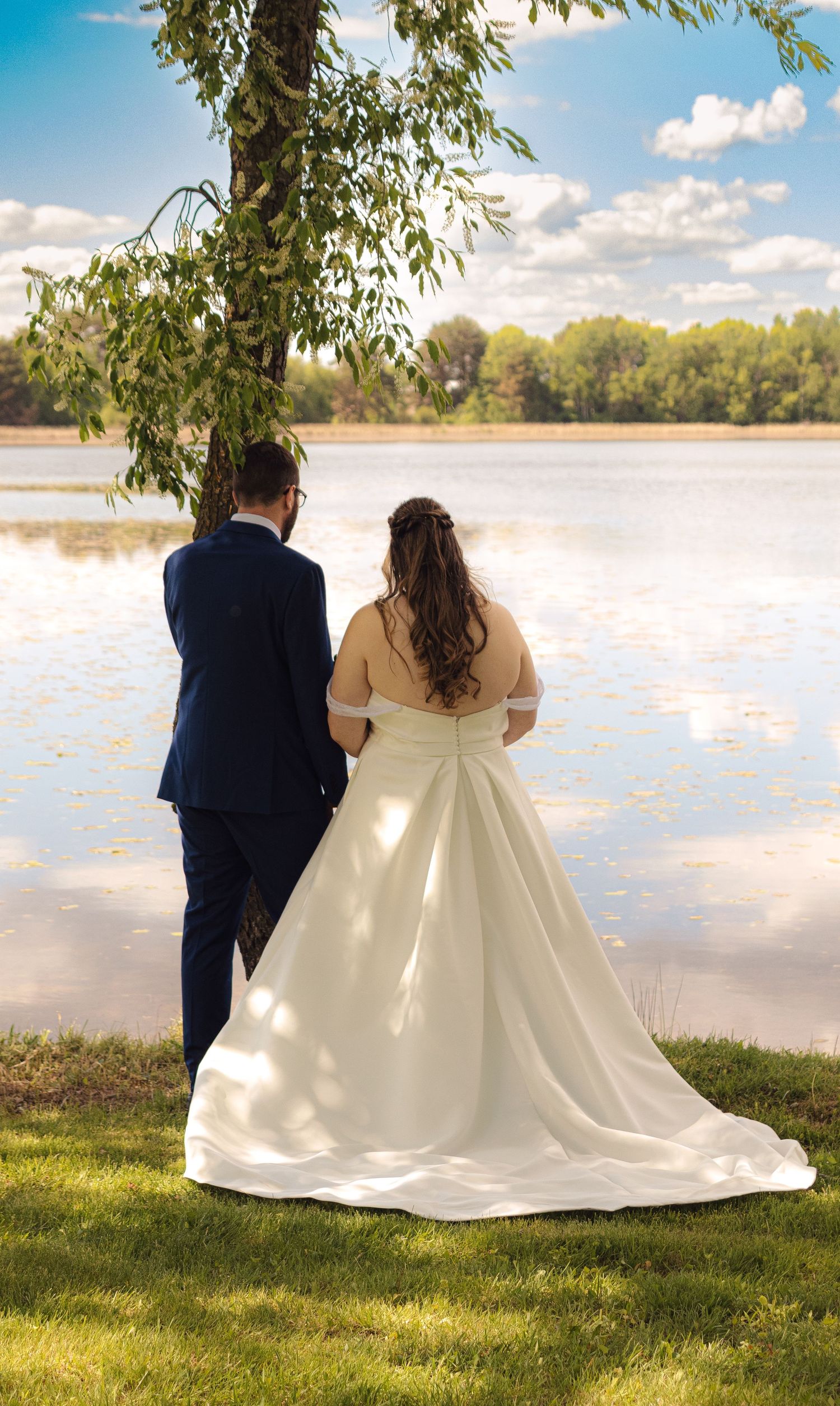 A romantic lakeside scene with a couple in wedding attire sitting on grass overlooking the water on a sunny day.