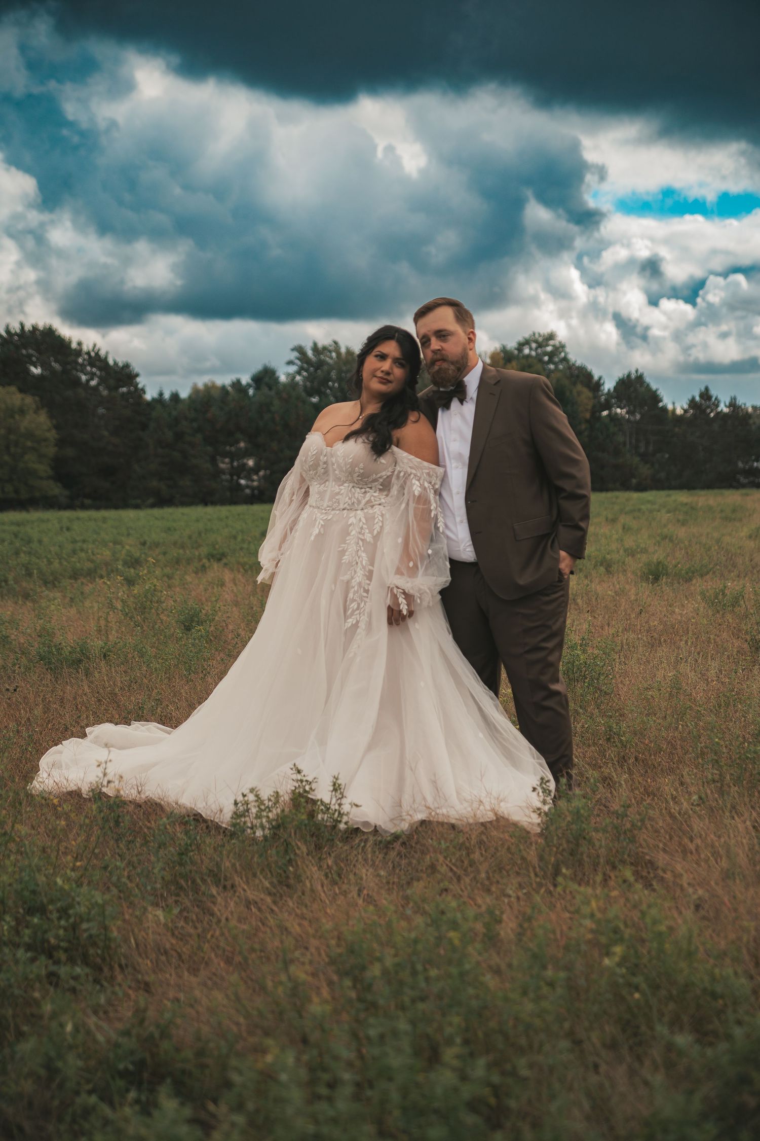 Bride in white off-shoulder gown and groom in brown suit pose in a green field under dramatic cloudy sky.