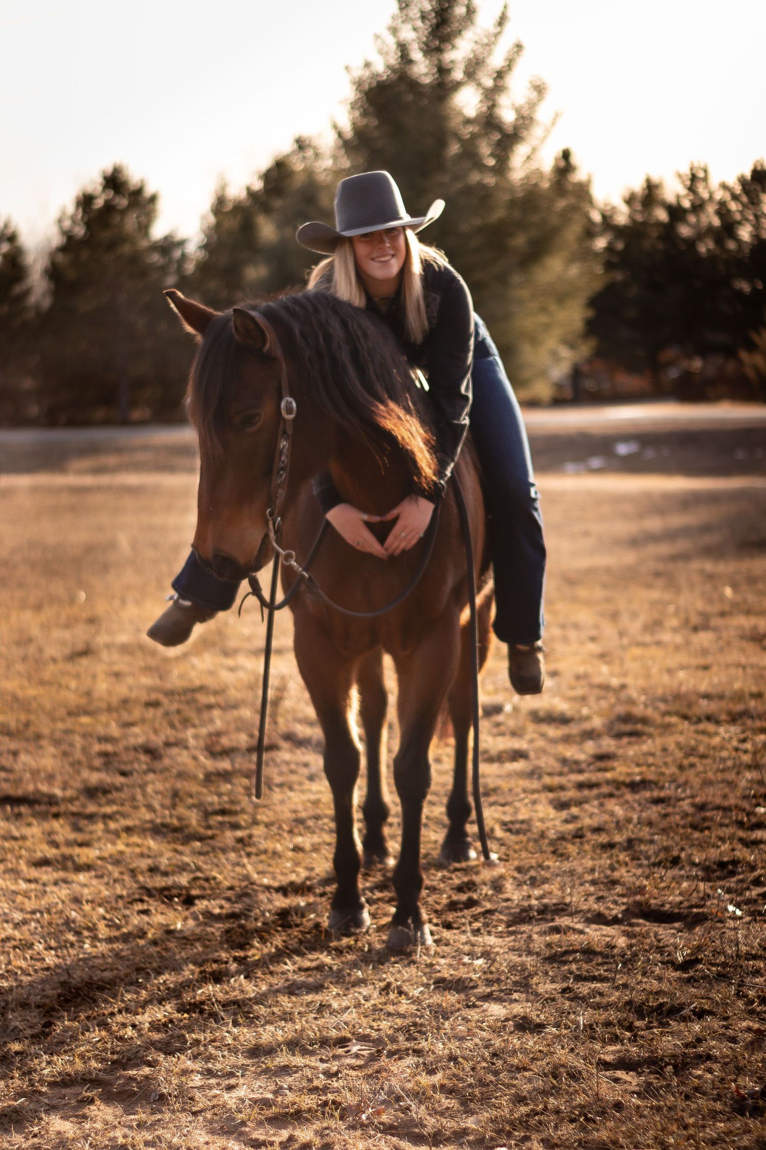 A cowboy-hat wearing figure walks alongside their horse in a sun-drenched field at dusk.