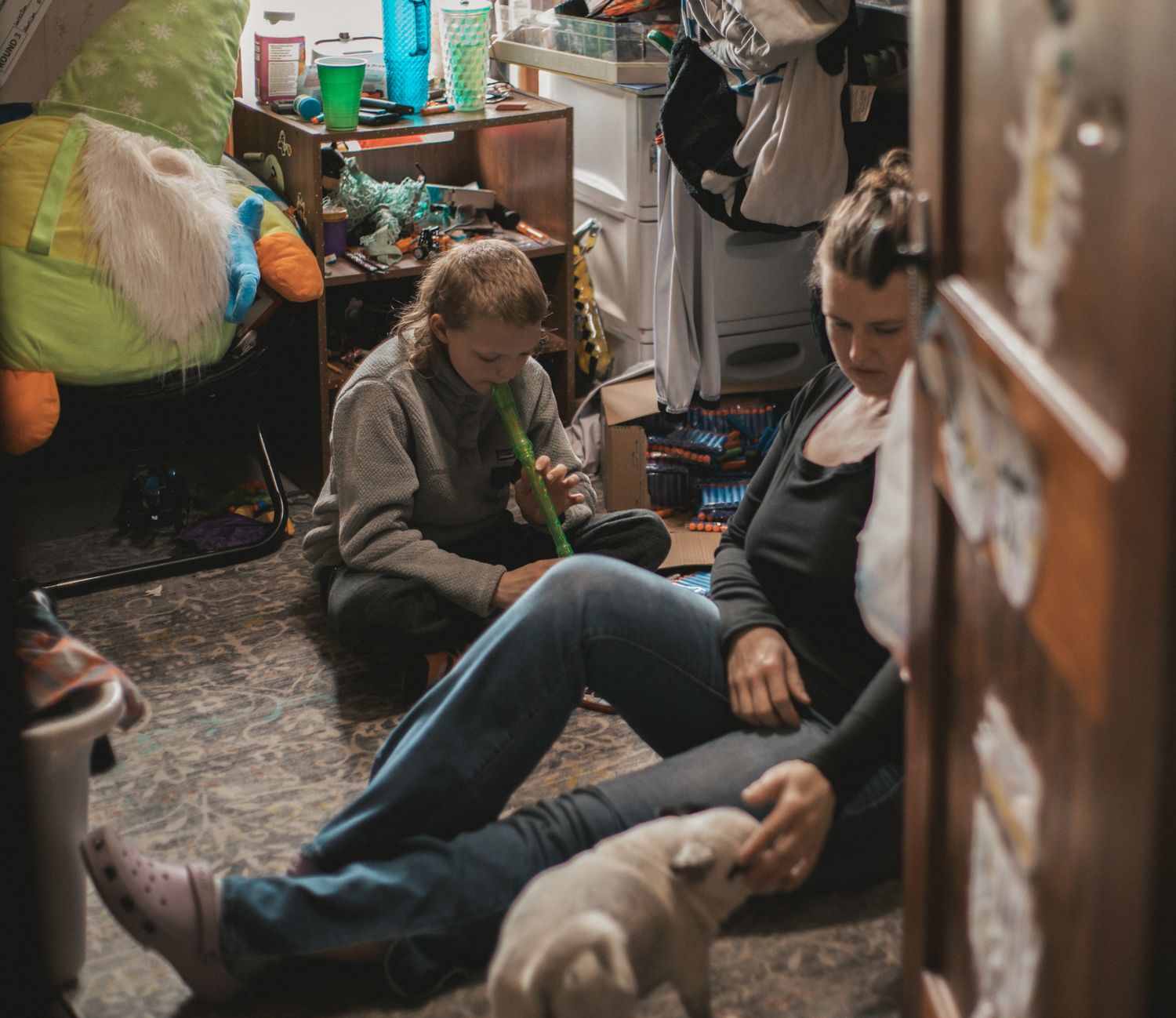 Two people sitting on a cluttered floor in a messy room surrounded by various items and belongings.