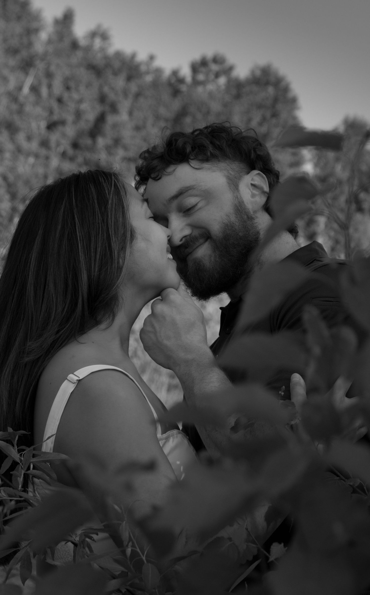 A romantic black and white portrait of a couple embracing among foliage in soft natural lighting.