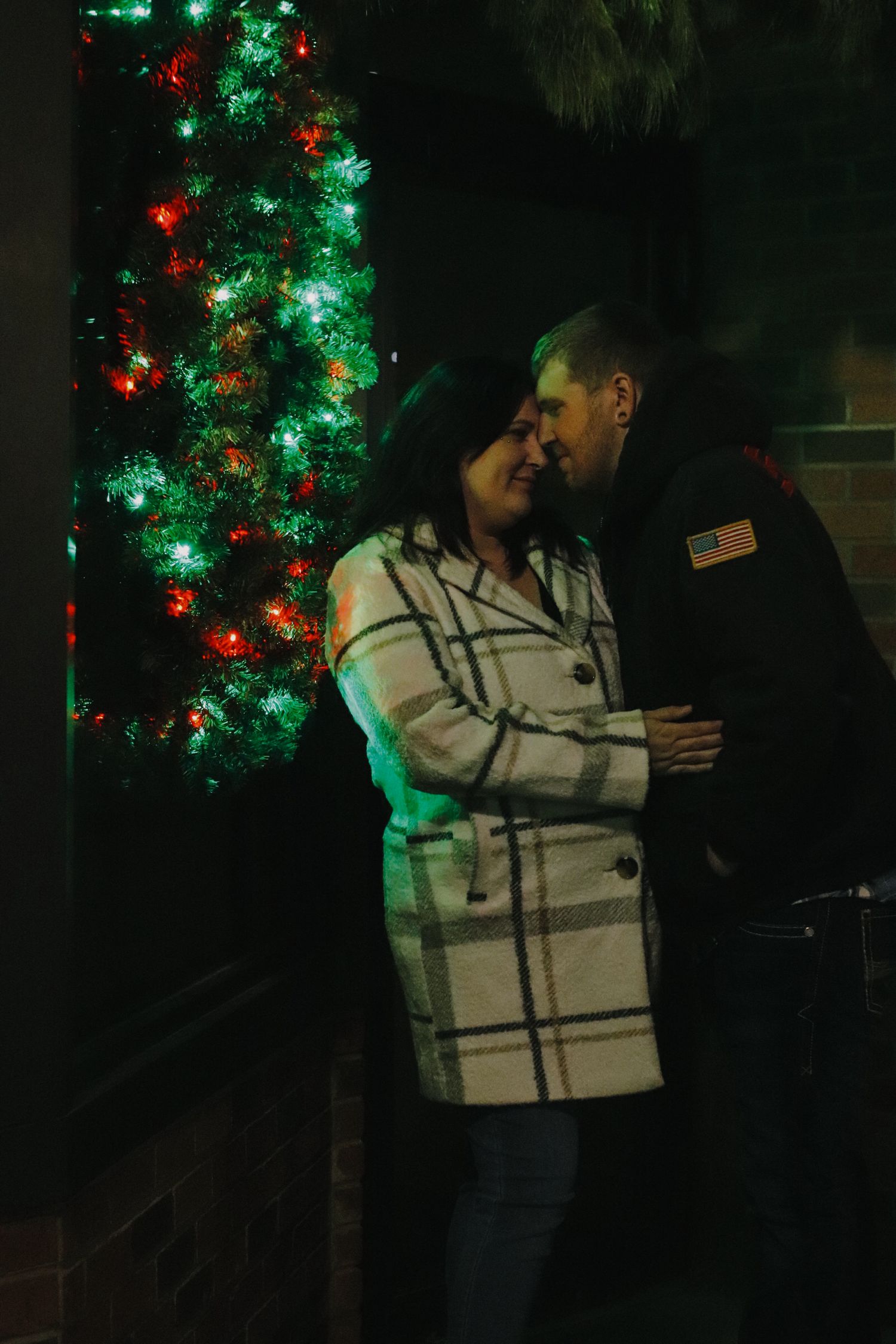 A person in a plaid sweater stands by a decorated Christmas tree with glowing green and red lights in the dark.